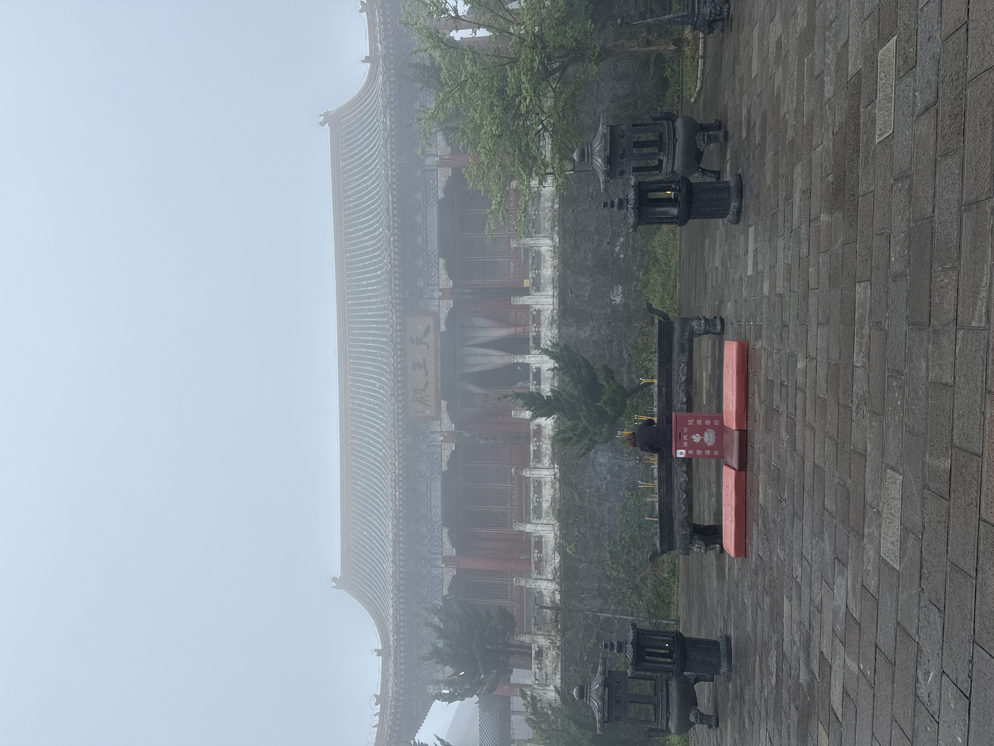 The image depicts a building, partially obscured by fog, in the Yongding District, China. The building has a traditional Chinese architectural style with a multi-tiered roof and intricate window designs. A person stands in front of a red rectangular structure with Chinese characters, possibly a donation box or offering table. Incense sticks are visible near the person. In the foreground and background are stone pavers and ornamental lanterns. The environment is overcast, with heavy fog impacting visibility.