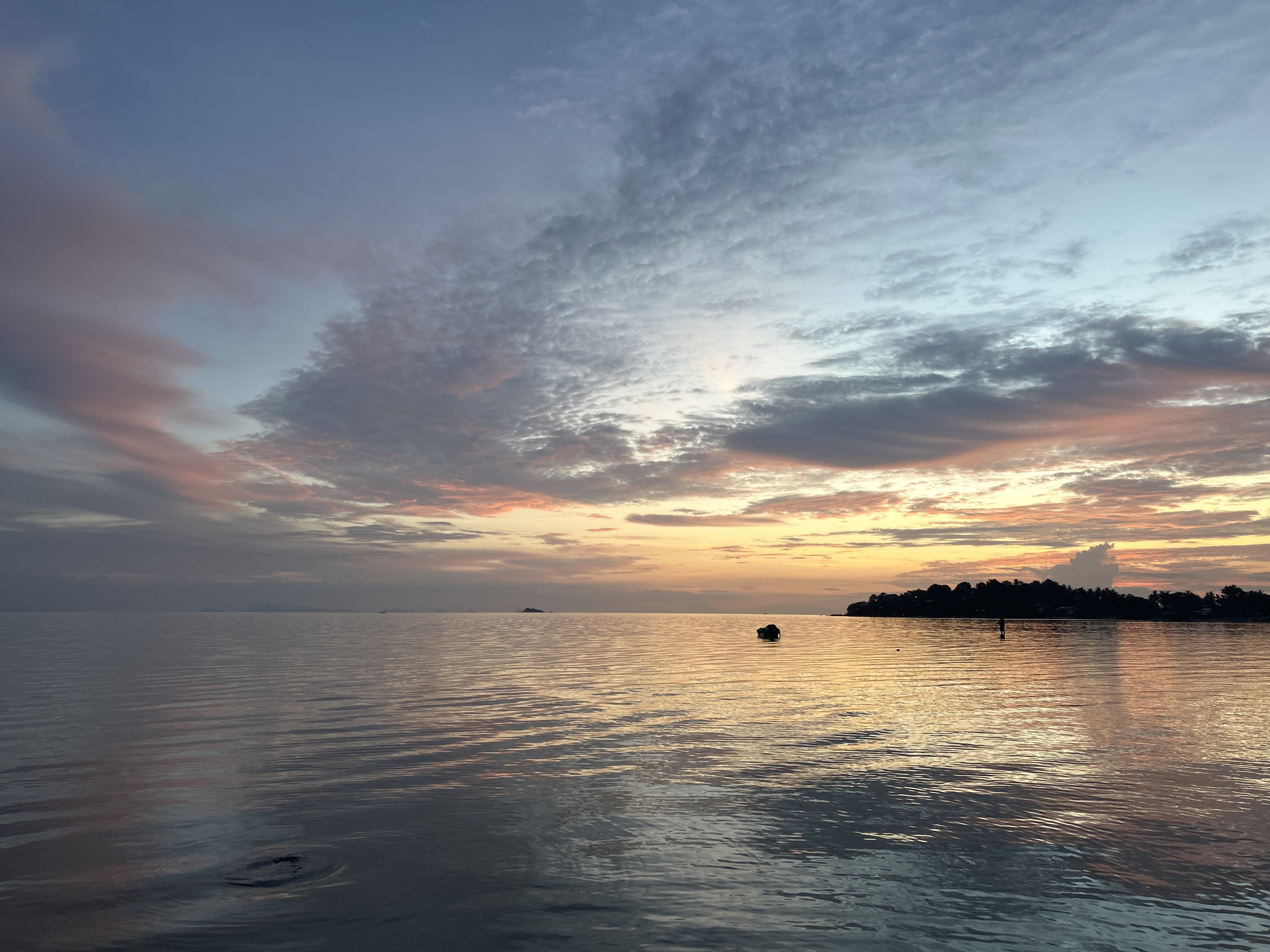 The image shows a serene seascape at sunset on Ko Pha-ngan District, Thailand. The sky is painted with hues of blue, pink, and orange, with wispy clouds scattered across it. The calm ocean reflects the colors of the sky, creating a shimmering effect on the water's surface. In the distance, a silhouette of land with trees and buildings is visible on the horizon, suggesting the presence of a coastline. A small, dark object, possibly a boat or a marker, floats on the water in the middle ground. The overall atmosphere is tranquil and picturesque, capturing the beauty of a tropical sunset.