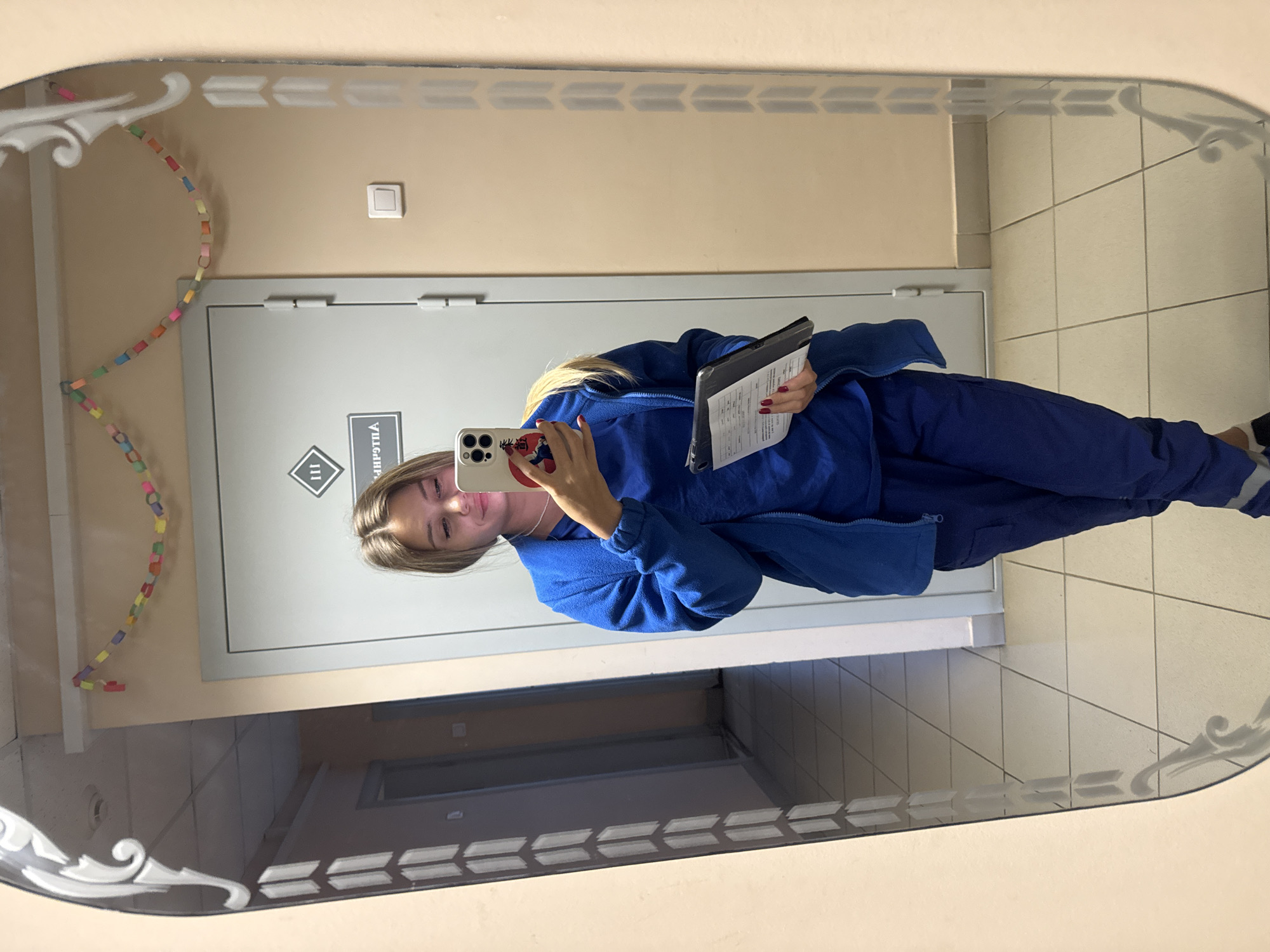 A young woman with blonde hair is taking a mirror selfie in a hallway. She is wearing blue scrubs, a blue zip-up jacket, and dark blue pants. She is holding a tablet in her left hand and a phone in her right hand, which she is using to take the picture. The hallway has light-colored walls and tiled floors. A closed door is visible behind her, with a sign above it that reads "III". A festive paper chain decoration is hanging on the wall above the door. The mirror has an ornate, decorative border. The overall scene suggests a medical facility or a similar institutional setting. The location is not definitively Chaikovsky, Russia based on the visual information provided.