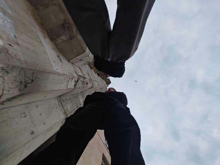 The image depicts a low-angle view upwards in Shumen, Bulgaria, capturing a partial architectural scene under an overcast sky. In the immediate foreground, the lower bodies of two individuals are visible. One person, positioned bottom-center and left, wears black pants. The other individual, whose legs in dark grey pants are seen top-right, extends downward into the frame. The camera perspective places the viewer directly beneath or between these subjects.
The left and central portions of the image are dominated by a tall, weathered concrete structure, likely a building wall or support pillar. This structure exhibits rough textures, visible seams, cracks, and patches of discoloration. Dark graffiti, consisting of abstract lines and symbols, is present on the lower section of the concrete surface. Further right, partially obscured by the person in black pants, a section of a lighter-colored building facade, possibly stone or brick, is visible, featuring a dark rectangular opening indicative of a window. The sky overhead is uniformly cloudy and light grey. A single small, dark bird is in flight in the upper-right quadrant of the sky.