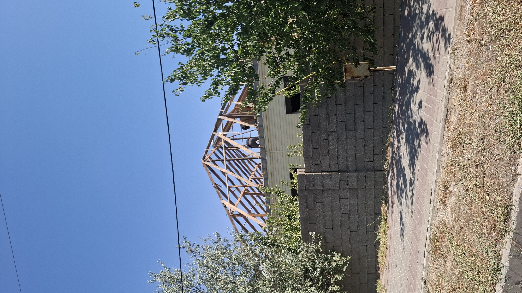 The image shows a house under construction in Shymkent, Kazakhstan. The roof is partially visible, with wooden beams forming the structure. Two individuals are working on the roof. A grey brick wall separates the property from a paved walkway. Trees with green foliage are present on either side of the house. The sky is clear and blue.
