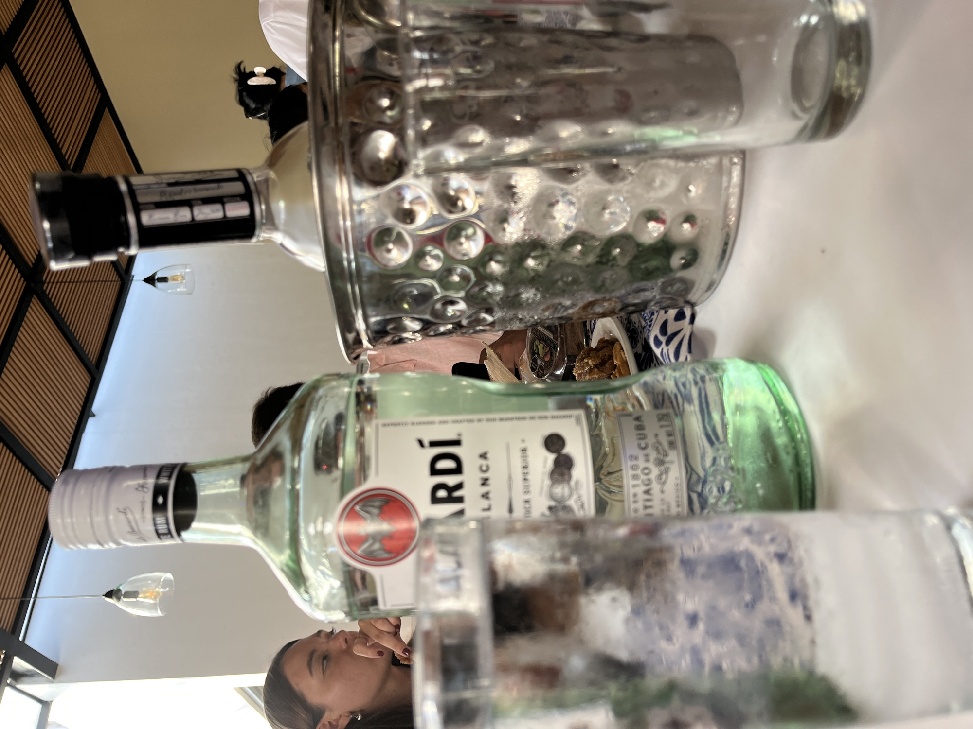 A photograph taken indoors shows a scene with multiple bottles and individuals. A bottle of Bacardi Blanca rum is prominently displayed, with its label clearly visible. Another bottle, inside a metallic ice bucket with a hammered texture, is present. A clear glass is positioned in the foreground, partially obscuring the view of people in the background. One individual, identifiable as a woman, is seen in the lower-left corner. The context suggests this scene might be in a restaurant or bar. The location provided, San Bernardino Tlaxcalancingo, Mexico, indicates the geographic setting of the scene.