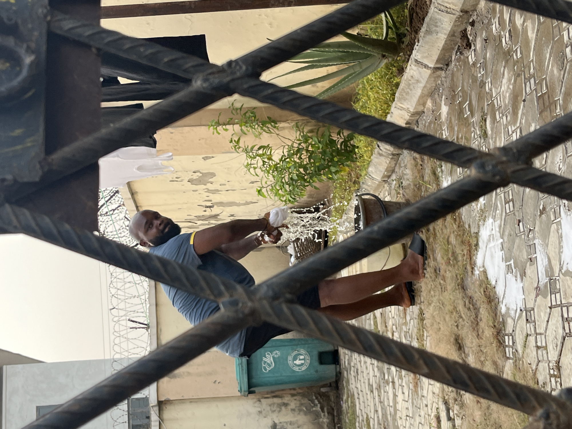 The image captures an outdoor scene, likely a yard or pathway, viewed through the blurry, dark, twisted bars of a metal gate or fence in the foreground. The primary subject is a bald, bearded man, wearing a blue short-sleeved shirt with a yellow stripe on the shoulder and dark shorts. He is barefoot, though a black slipper is partially on one foot.
The man is actively wringing out a white cloth or towel over a dark bucket, causing water to splash outward. The ground around him is paved with patterned tiles, which appear wet in several spots. Patches of green grass and some plants, including a large agave, are visible around the paved area.
In the mid-ground, a green plastic waste bin is situated, featuring a circular white logo. The logo depicts hands dropping waste into a bin and contains visible text, including "CLEAN ENVIRONMENT" and "KEEP." The background reveals a building wall and a fence topped with coiled barbed wire, suggesting a secure or residential area in Dushepe, Nigeria. The lighting is bright and even, indicating it is daytime. The man's expression appears neutral, focused on the task of wringing out the cloth.