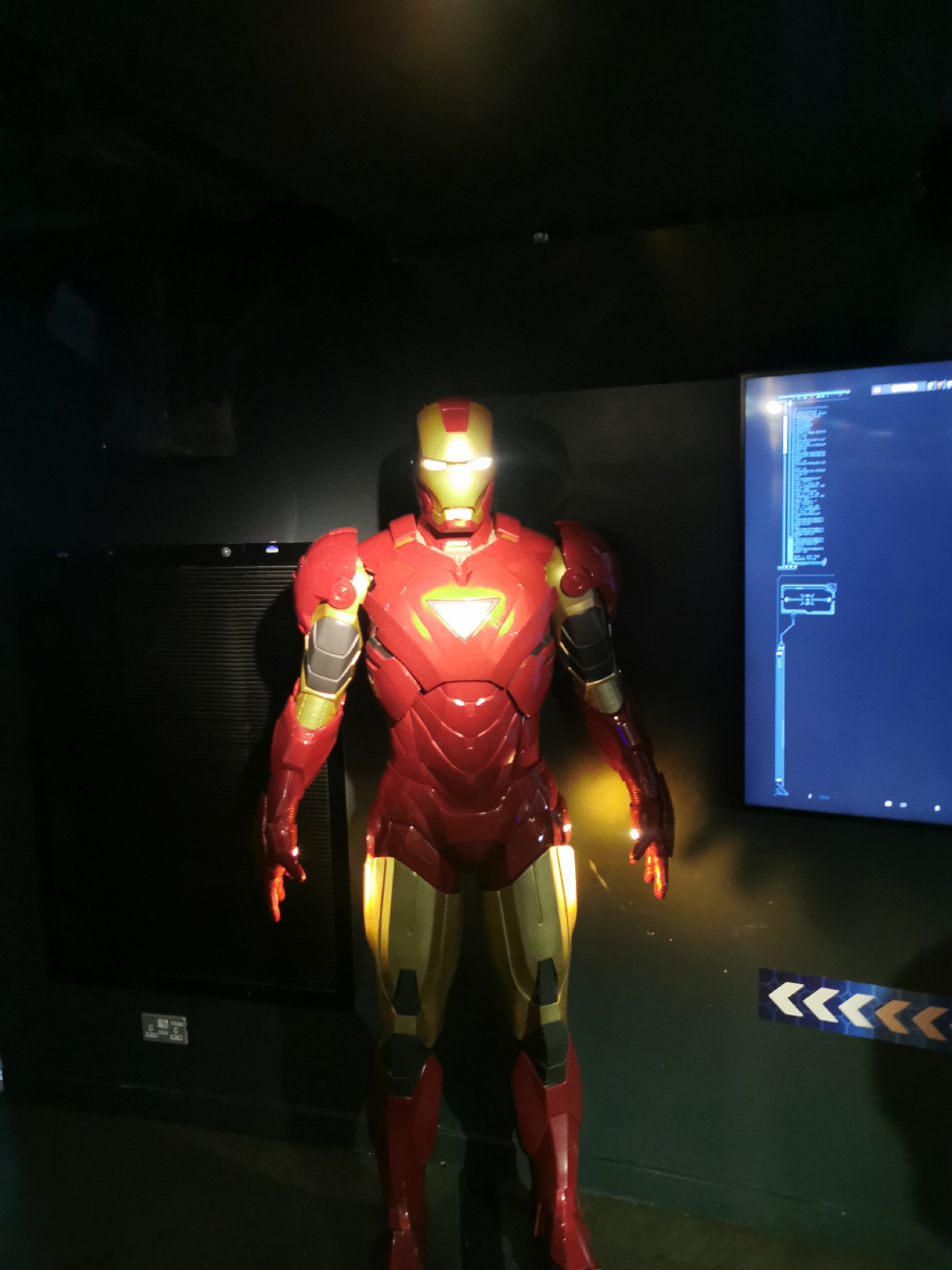 The image shows an Iron Man figure in London, United Kingdom. The figure is red and gold and is standing in front of a black wall. To the right of the figure is a television screen displaying data. At the bottom right of the image, there is a piece of tape with arrows printed on it.