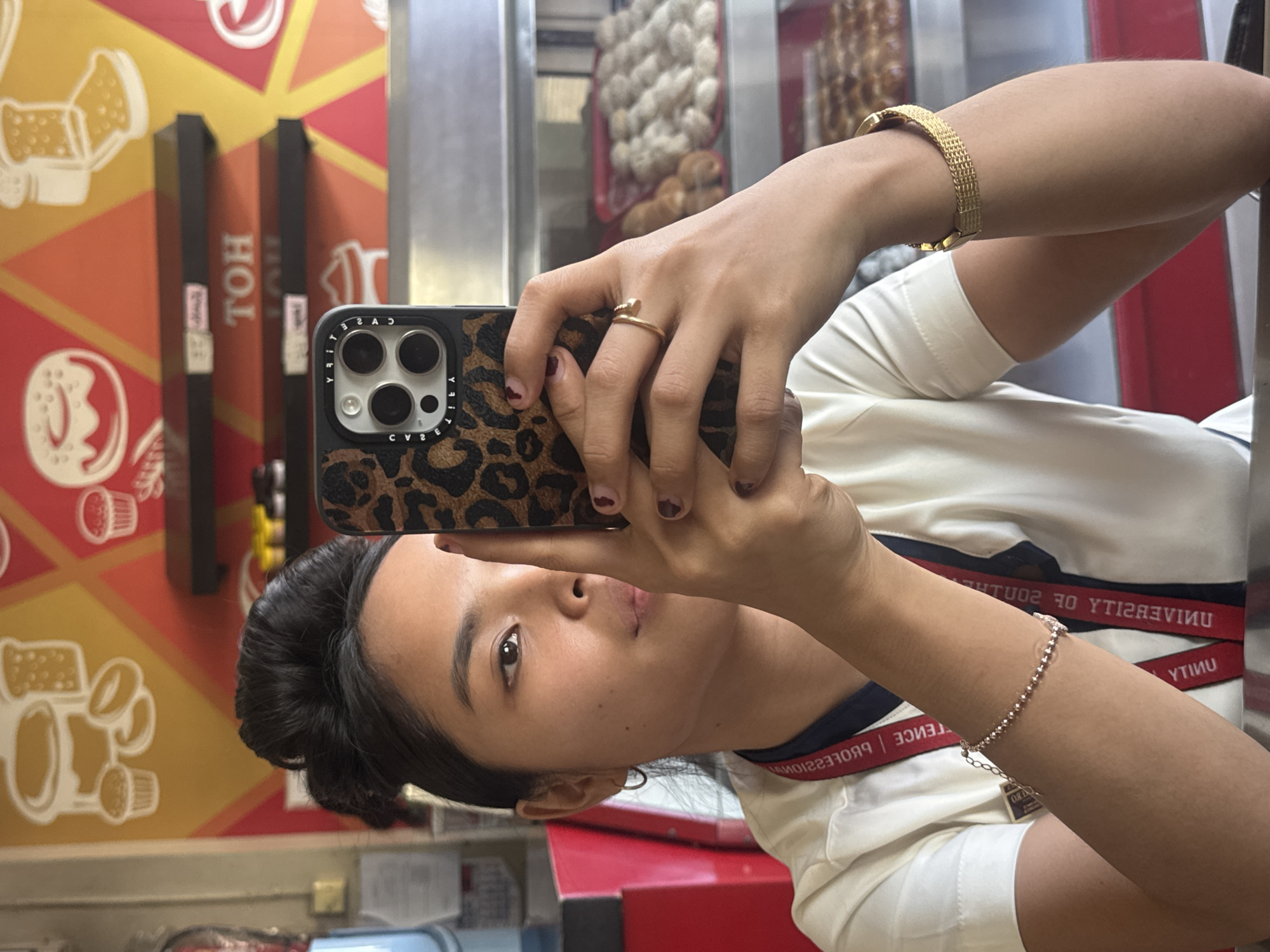 In an indoor establishment resembling a bakery or cafe in Davao City, Philippines, a young woman with dark hair styled in a bun is taking a selfie. She is wearing a white collared shirt with a navy blue undershirt and a red lanyard with white text. She has a silver bracelet on her left wrist and a gold bracelet on her right wrist, along with a gold ring on her right index finger. The background features a colorful mural depicting baked goods and shelving units containing pastries. The woman is holding a smartphone with a leopard-print case and a prominent camera module. The scene appears to be a casual moment captured within a commercial setting.