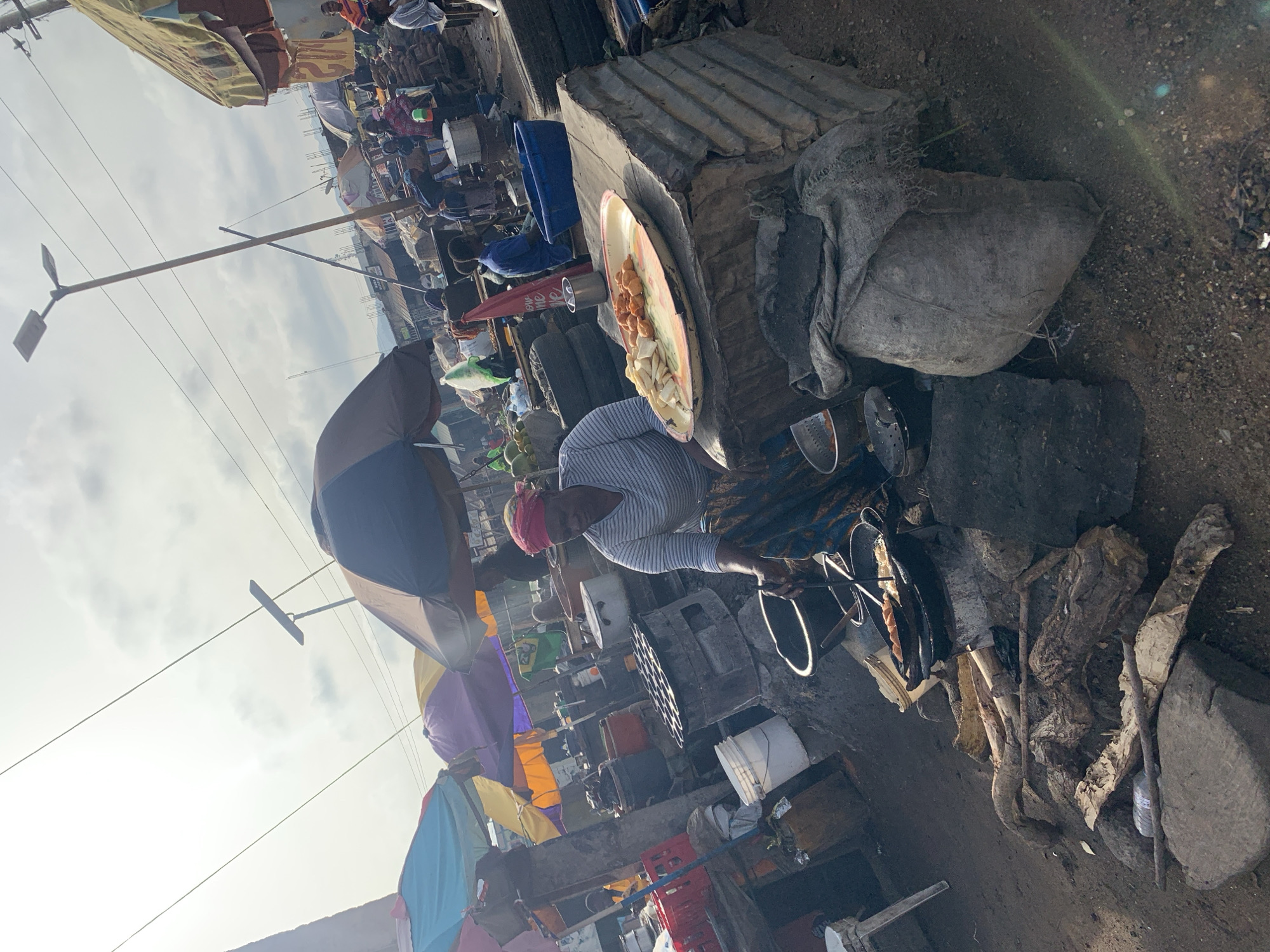This image captures a vibrant outdoor market scene in Rafin Par, Nigeria, teeming with daily activity. In the foreground, a person, identifiable by a red headscarf and striped shirt, is actively engaged in cooking or preparing food. They are bent over an open-fire cooking setup, where pans or pots are being heated. Logs and charcoal are visible on the dirt ground as fuel.
To the right of the cook is a makeshift counter or table fashioned from corrugated metal sheeting, on which rests a large, round, light-colored tray filled with food items. These include several golden-brown, fried-looking pieces (possibly akara or puff-puff) and some white, sliced pieces that could be yam or potato. Various cooking utensils, such as metal bowls and strainers, are scattered around the cooking area. A large, dark-colored umbrella provides shade for the cook, suggesting strong sunlight.
The background reveals a bustling market environment with numerous other stalls and vendors. Colorful umbrellas and canopies in shades of blue, yellow, and purple are visible, providing shade for other vendors and their wares. Distant figures can be seen moving about, adding to the sense of a lively marketplace. Power lines and a street light pole stretch across the bright, somewhat hazy sky, which suggests it is daytime, likely mid-morning or afternoon, given the intensity of the light. The overall setting is characterized by informal commerce and resourceful, makeshift structures on a dirt ground. No clearly legible text is visible within the frame. The scene conveys a sense of industriousness and community daily life.