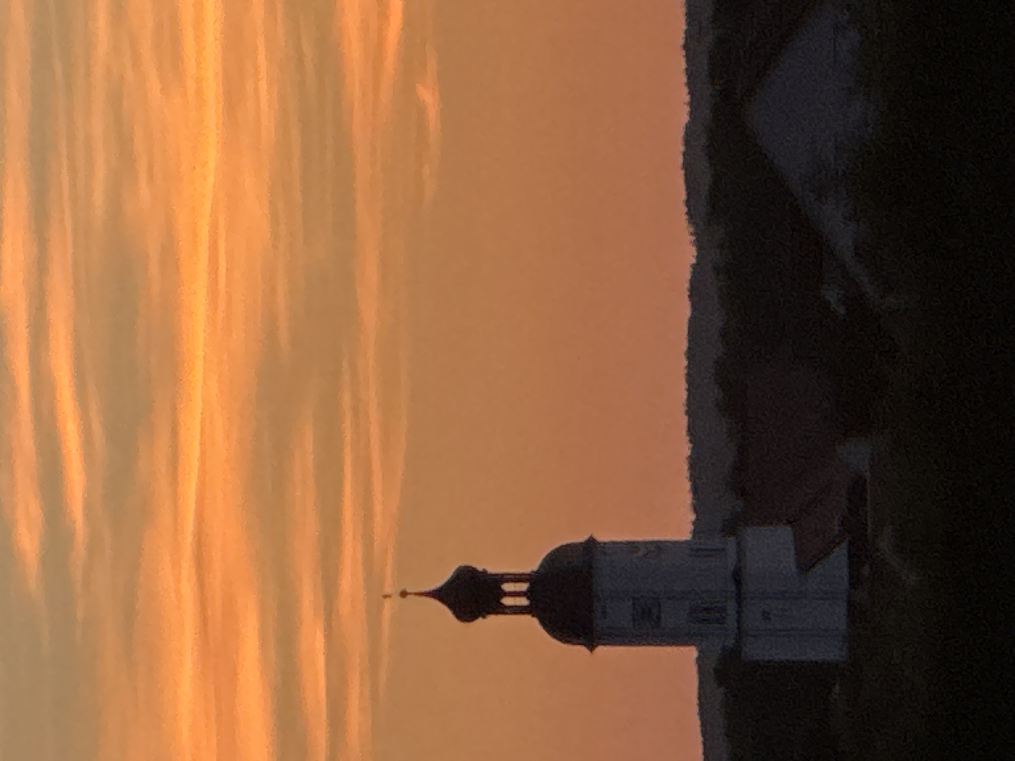 This image depicts a church steeple silhouetted against a vibrant orange and yellow sunset sky. The sky is filled with horizontal, textured clouds that are illuminated by the setting sun. Below the sky, a dark, silhouetted landscape with trees and buildings is visible. The prominent structure is a white church tower with a dark, onion-shaped dome and a cross on top. The tower has visible windows and architectural details. To the right of the church, part of a dark, sloped roof of another building is visible. The overall scene is one of peaceful evening light over a town. The church steeple is likely St. Georg, a landmark in Wettstetten, Germany.