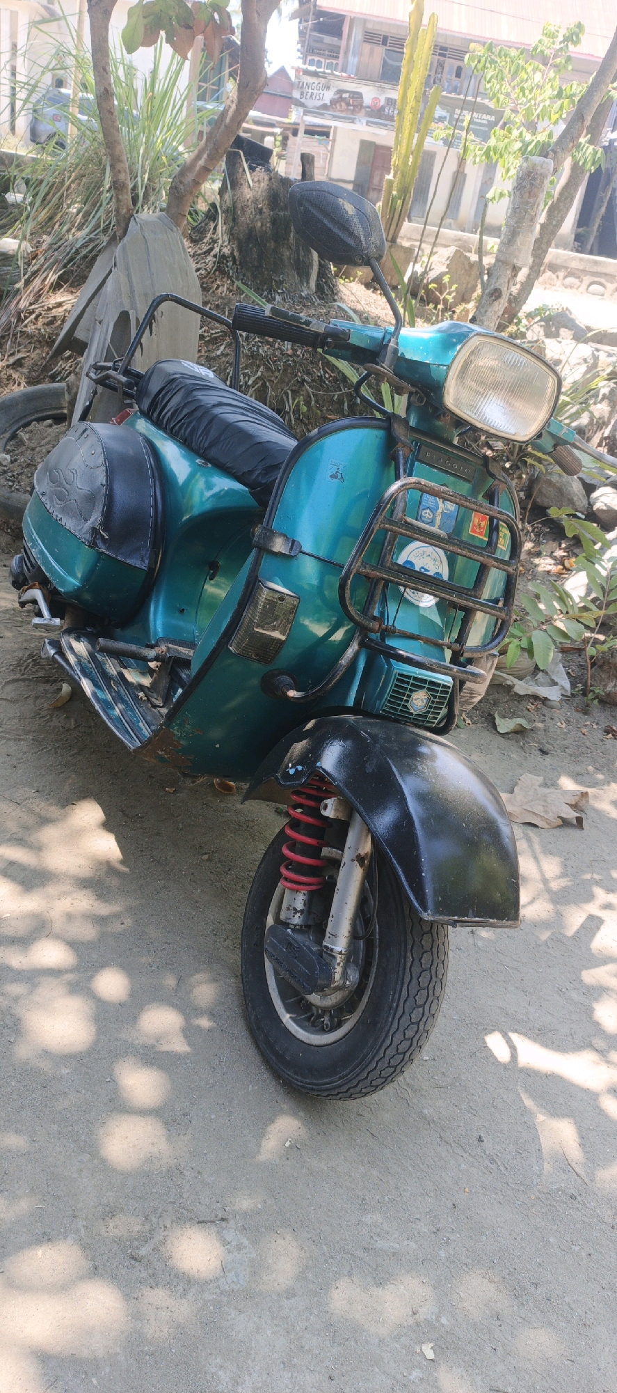 The image shows a turquoise vintage scooter parked outdoors on a dirt or gravel surface. The scooter has a black seat, a black front fender, and a black luggage rack on the front. Red springs are visible on the front suspension. The background includes trees, vegetation, and buildings that appear to be in a rural or semi-urban setting, consistent with a location in Sidodadi, Indonesia. The lighting suggests it is daytime, with dappled sunlight casting shadows on the ground. There are no people visible in the image.