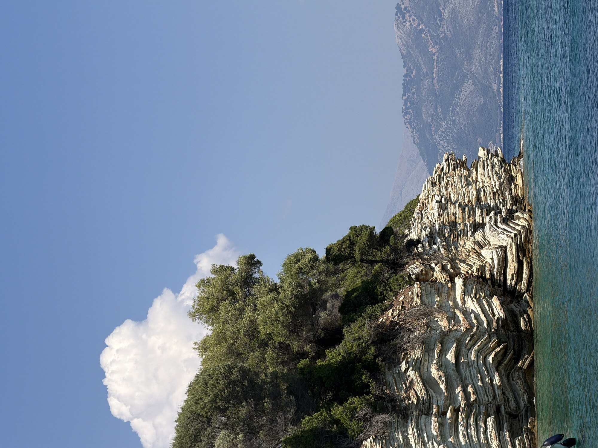 A picturesque scene unfolds in the Municipal Unit of Kastos, Greece, where a rocky coastline meets the clear blue sea under a vast, cloud-speckled sky. The foreground is dominated by a stratified rock formation, its layers sculpted by time and tide into undulating patterns. Lush green vegetation, including trees and shrubs, clings to the top of the cliff, their branches reaching out towards the sky.
In the distance, hazy mountains rise, their slopes dotted with sparse vegetation, providing a soft backdrop to the vibrant blues of the sea and sky. The water itself is a deep, inviting turquoise, its surface gently rippled. A sliver of a black and white object, possibly a buoy or fender, is visible in the lower left corner, hinting at the presence of a vessel nearby. The overall impression is one of natural beauty and tranquility, a serene snapshot of a coastal landscape.
