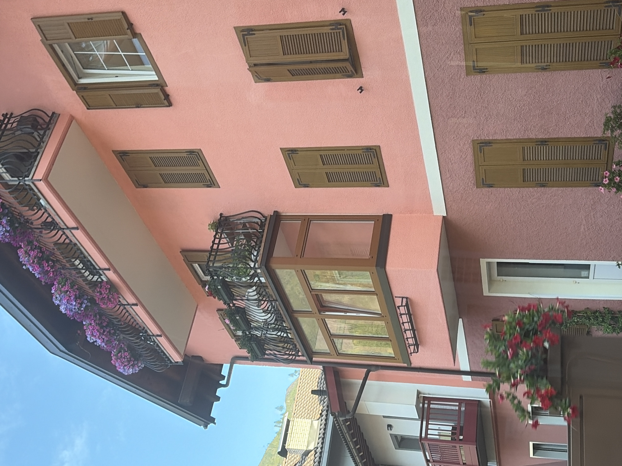 A close-up, side-angle view of a multi-story building in Predazzo, Italy, displays a vibrant pink facade adorned with numerous wooden shutters. The building features multiple balconies, one with a black wrought-iron railing overflowing with pink and purple flowers. Another, smaller balcony with a red railing is visible on an adjacent section of the building. A prominent bay window with wooden frames interrupts the facade. The sky above is clear and blue, and a hint of a mountain landscape can be seen in the background. A planter box with red flowers is positioned in the foreground.