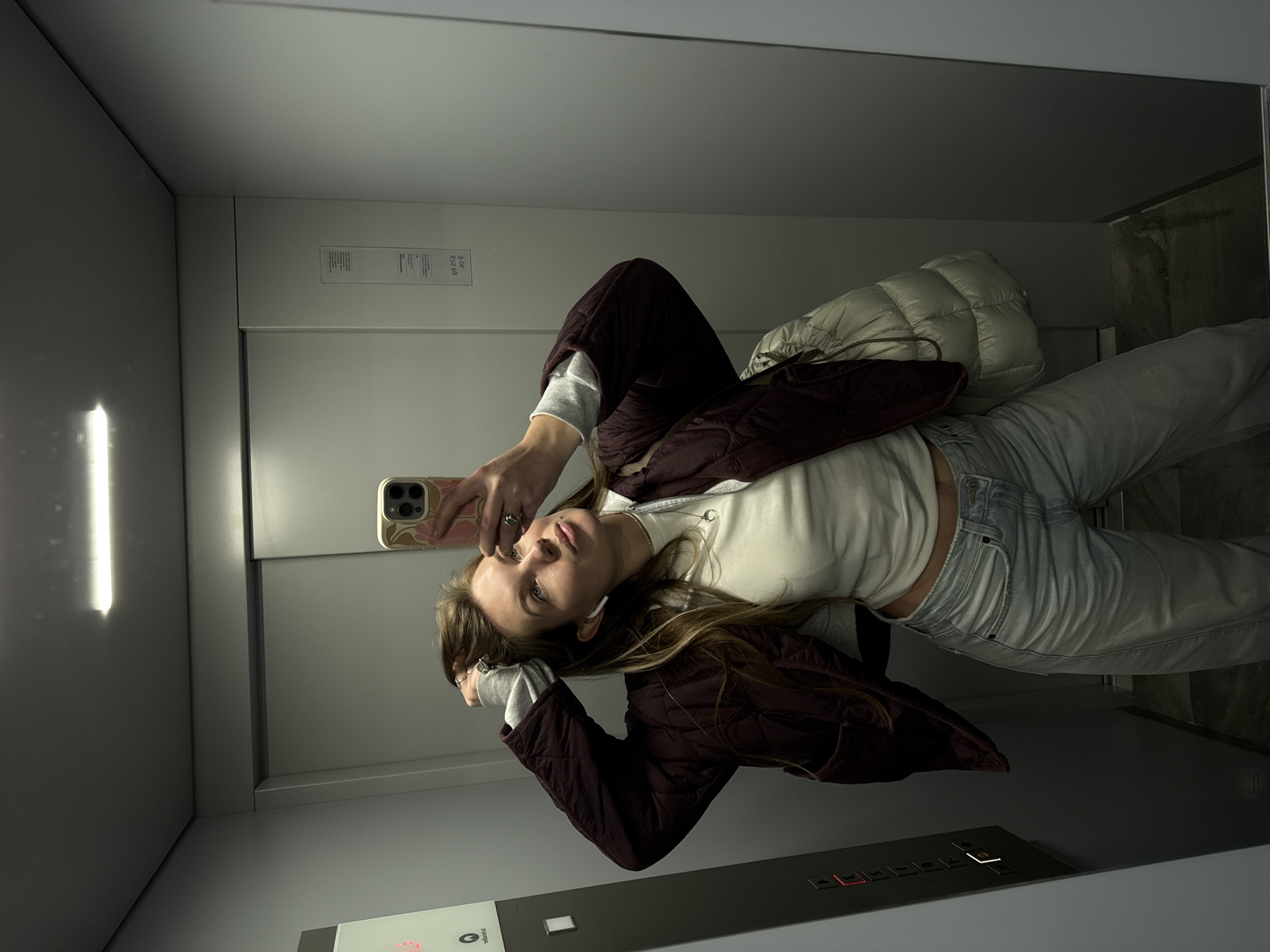 A young woman is taking a selfie in an elevator. She is wearing a white long-sleeved shirt, a burgundy quilted jacket, and light-wash jeans. She is holding a smartphone in her right hand, and her left hand is near her head. She also has a light-colored quilted bag slung over her shoulder. The elevator has a light panel on the ceiling and a control panel with illuminated buttons. The setting appears to be within Vozdovac Urban Municipality, Serbia, given the user's prompt.