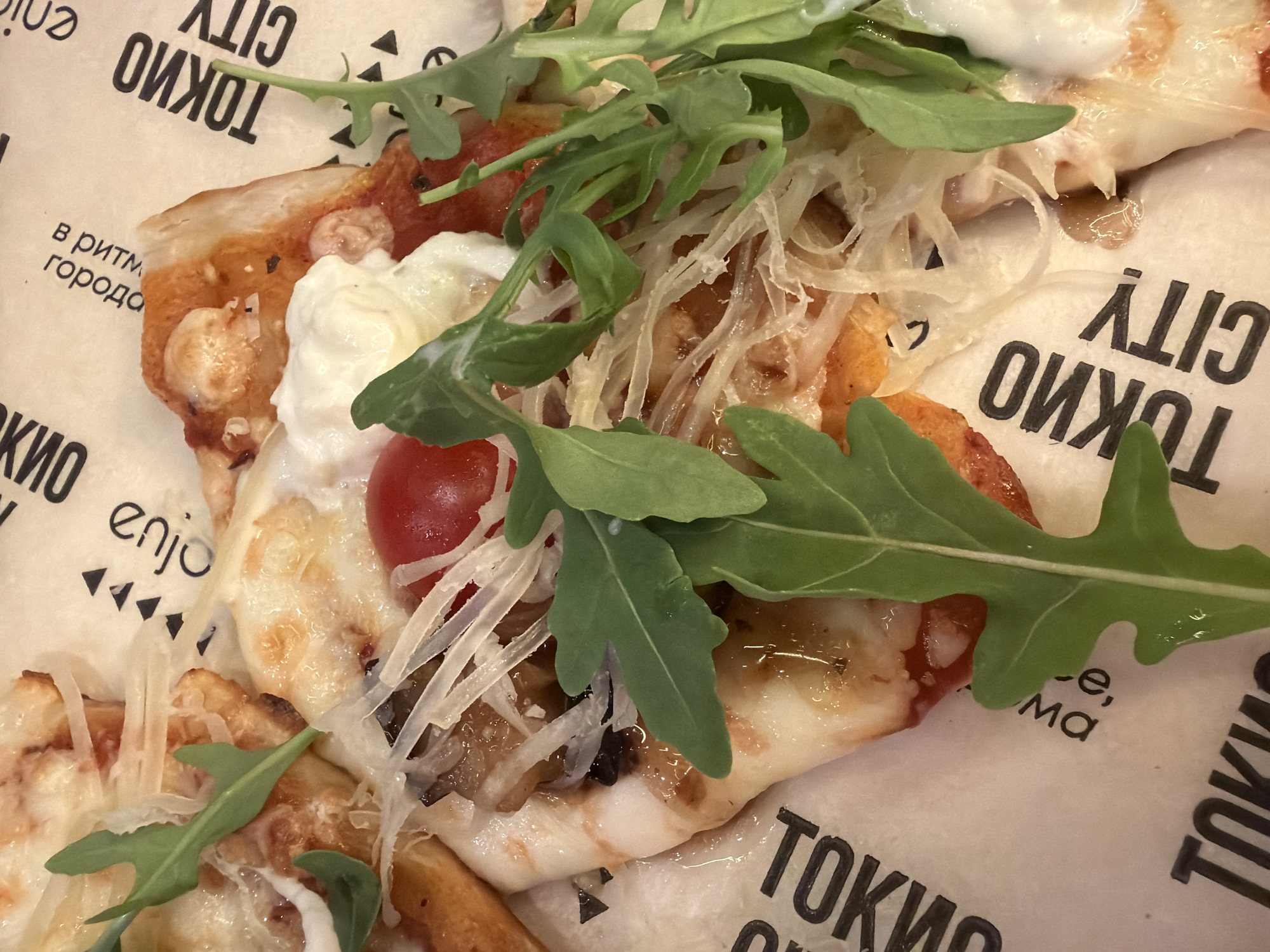 A close-up, overhead shot shows a slice of pizza placed on paper that reads "TOKYO CITY" in Russian. The pizza is topped with arugula, shredded cheese, and a cherry tomato. The crust is golden brown and appears to be thin. The paper has a black, repeating pattern of the words "TOKYO CITY" and small triangular shapes. In the background, out of focus, are additional pieces of pizza and the same printed paper.