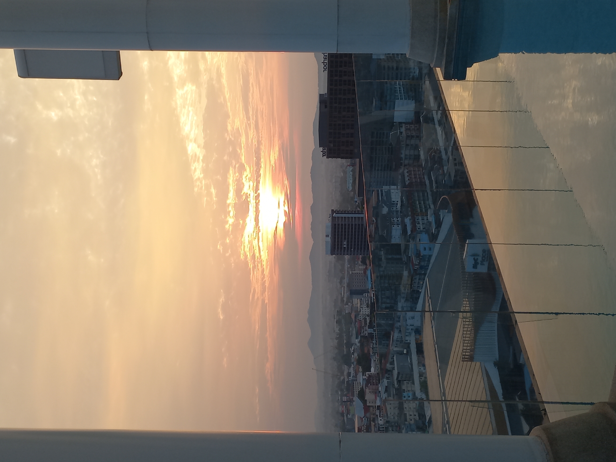 The image presents a sideways view of Pattaya City, Thailand, observed from an elevated perspective, possibly a rooftop or high-rise building. The time of day appears to be during sunset, as indicated by the warm, golden hues in the sky. The sun is partially obscured by clouds, creating a dramatic effect.
The city skyline includes a mix of high-rise buildings and lower-rise structures, suggesting a densely populated urban area. In the middle distance, there are hints of hills or mountains, indicating the city's geographical context.
In the foreground, parts of the observation point are visible, including structural elements that appear to be architectural or engineering components of the building. The composition gives the impression of looking out and over Pattaya from an elevated and enclosed space.