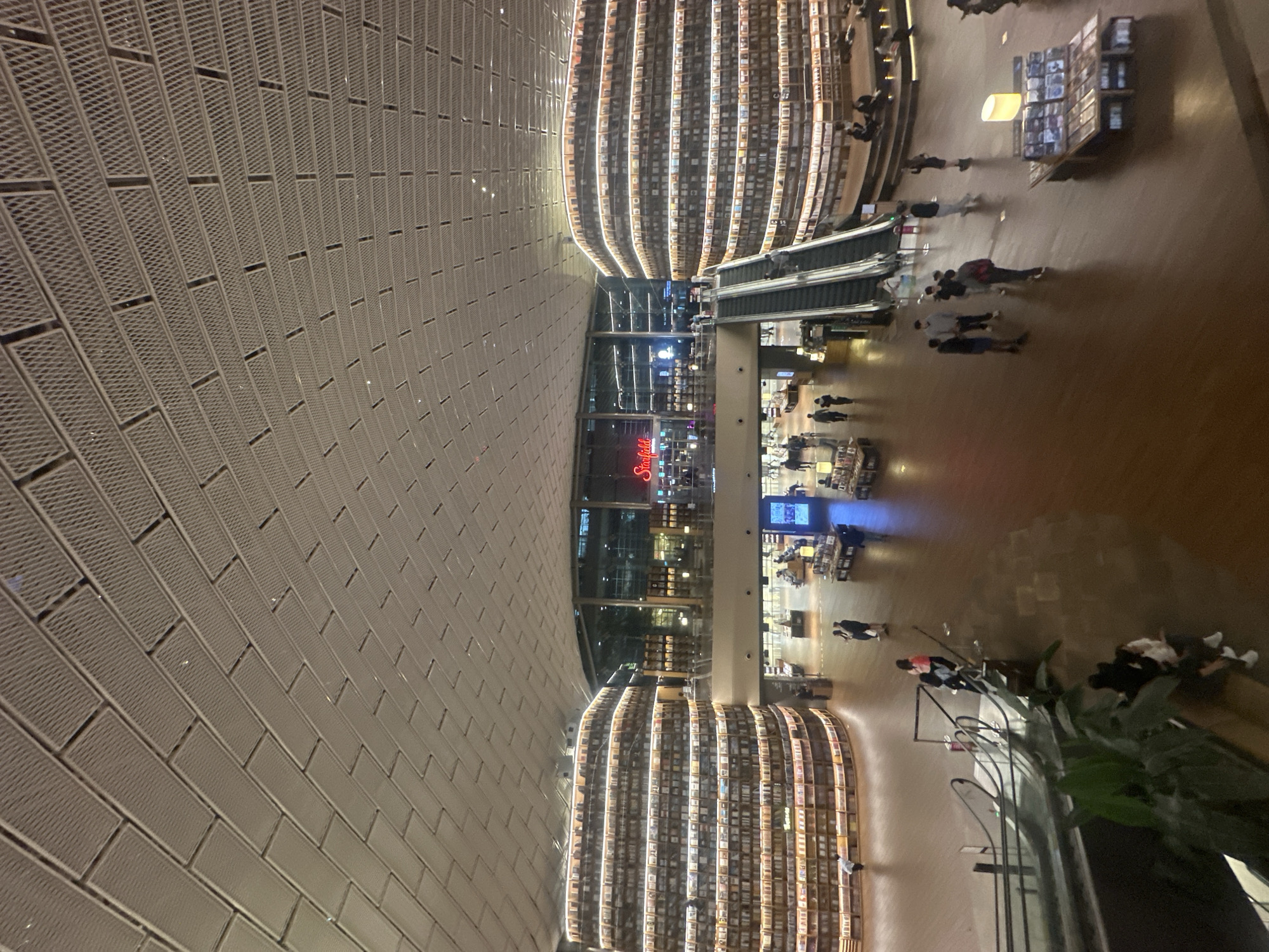 The image depicts the Starfield Library, located within the COEX Mall in Seoul, South Korea. The library features floor-to-ceiling bookshelves arranged in curved rows that appear to be a major architectural element. The building has a light grey ceiling with a grid pattern. Several escalators are visible in the center, leading to different levels of the library. Numerous people are seen walking around, browsing the shelves, or standing near escalators. There are some plants at the bottom right, in the foreground of the image. A "Starfield" sign is visible in the center of the photo. The photo is taken from a high vantage point, looking down into the library space.