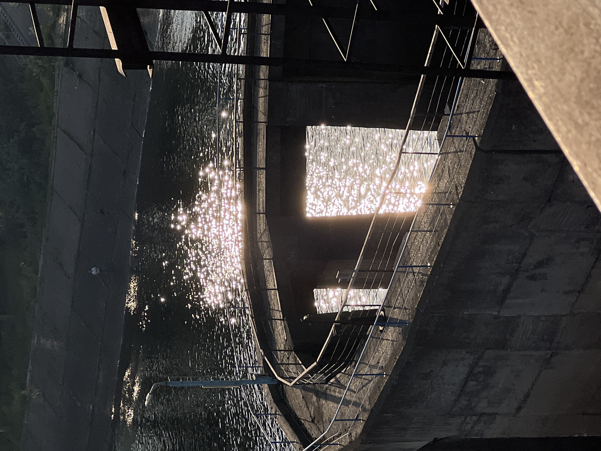 The image depicts a concrete dam structure with a curved walkway bordering a body of water. The water reflects sunlight, creating a shimmering effect across its surface. A solitary person is visible on the far bank of the water, appearing to be fishing. The walkway is equipped with blue-railed metal fencing. The overall scene is characterized by the interplay of concrete, water, and sunlight, suggesting a daylight setting. This structure is part of the Kama Hydroelectric Power Station located in Chaikovsky, Russia.
