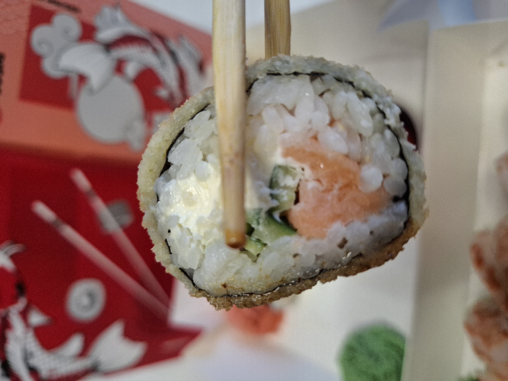 A close-up, slightly blurred shot shows a piece of sushi, likely a roll, being held by wooden chopsticks. The sushi appears to be deep-fried, with a crispy, golden-brown outer layer. Inside, layers of white rice, a pink filling (likely salmon), green cucumber, and a white creamy ingredient (possibly cream cheese or mayonnaise) are visible. In the background, a red container with white Asian-inspired motifs is partially visible. To the left and bottom of the sushi, out-of-focus green and pinkish elements suggest other food items, possibly wasabi and pickled ginger, or other sushi components, on a light-colored plate or surface. The lighting is bright, highlighting the textures of the food. The scene is devoid of any discernible landmarks or elements that would allow for the identification of a specific location within Bratislava, Slovakia.