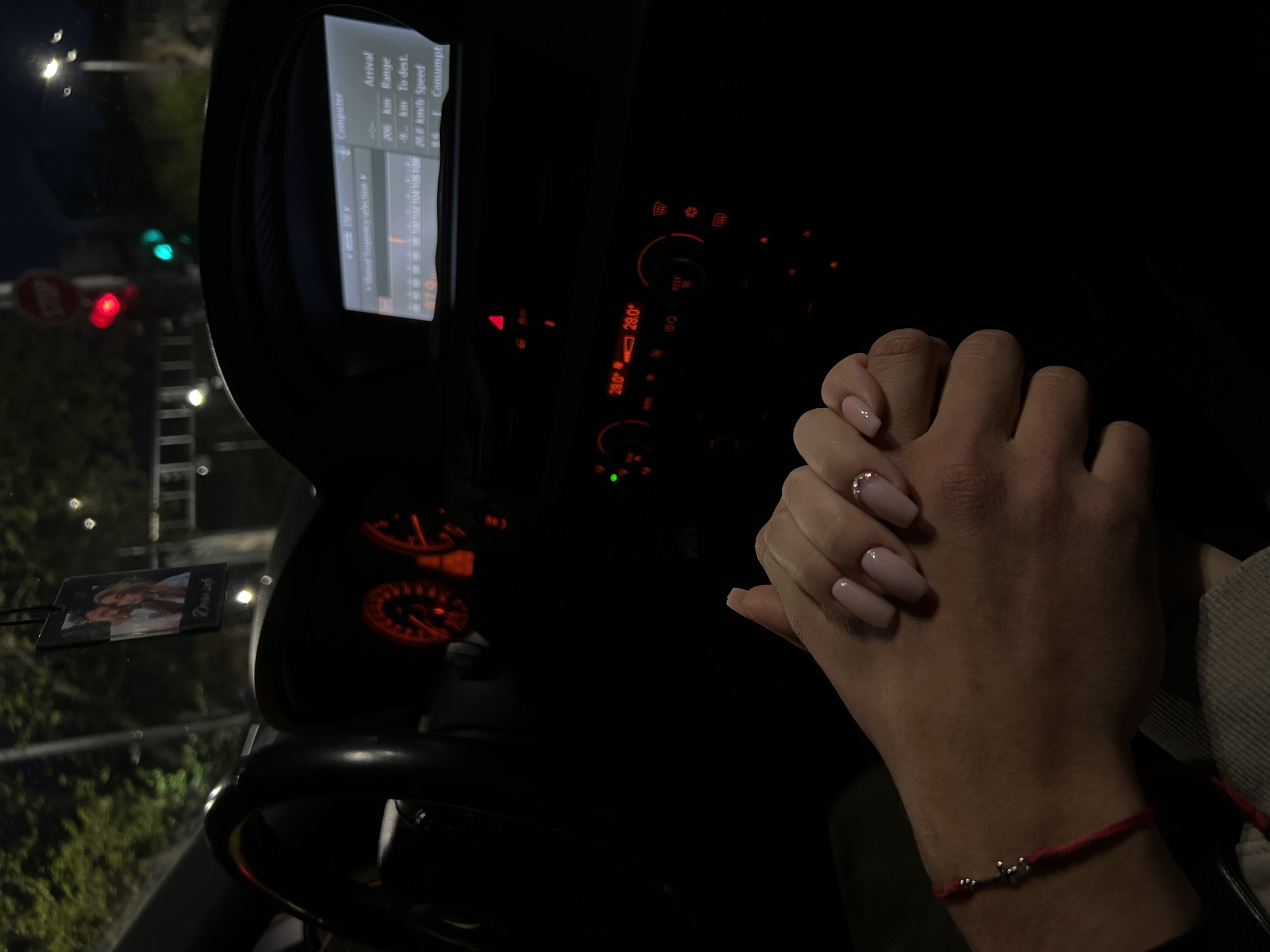 Inside a car at night in Plovdiv, Bulgaria, two individuals are holding hands, conveying intimacy and affection. The scene is illuminated by the car's interior lights and ambient street lighting.
The hands are clasped in the foreground, with one hand displaying light pink manicured nails, some adorned with small rhinestones, holding the other hand which wears a red string bracelet with a small silver cross-like charm.
Through the windshield, a red traffic light and a green traffic light are visible, along with a partially obscured "STOP" sign, suggesting the vehicle is stopped at an intersection. Streetlights and trees are also vaguely visible in the dark background.
The car's interior features a dashboard with glowing red gauges and a central console displaying "28.0" twice, likely indicating the cabin temperature, along with a "REST" button and hazard light control. An infotainment screen provides information from the "Computer," including "Arrival," "206 km Range," "km to dest." (kilometers to destination), "28.0 km/h Speed," and "Consumpt." It also shows "Manual frequency selection" and radio frequencies "102 104 106 108." A "DreamGirl" air freshener, featuring a photo of a woman, hangs from the rearview mirror.