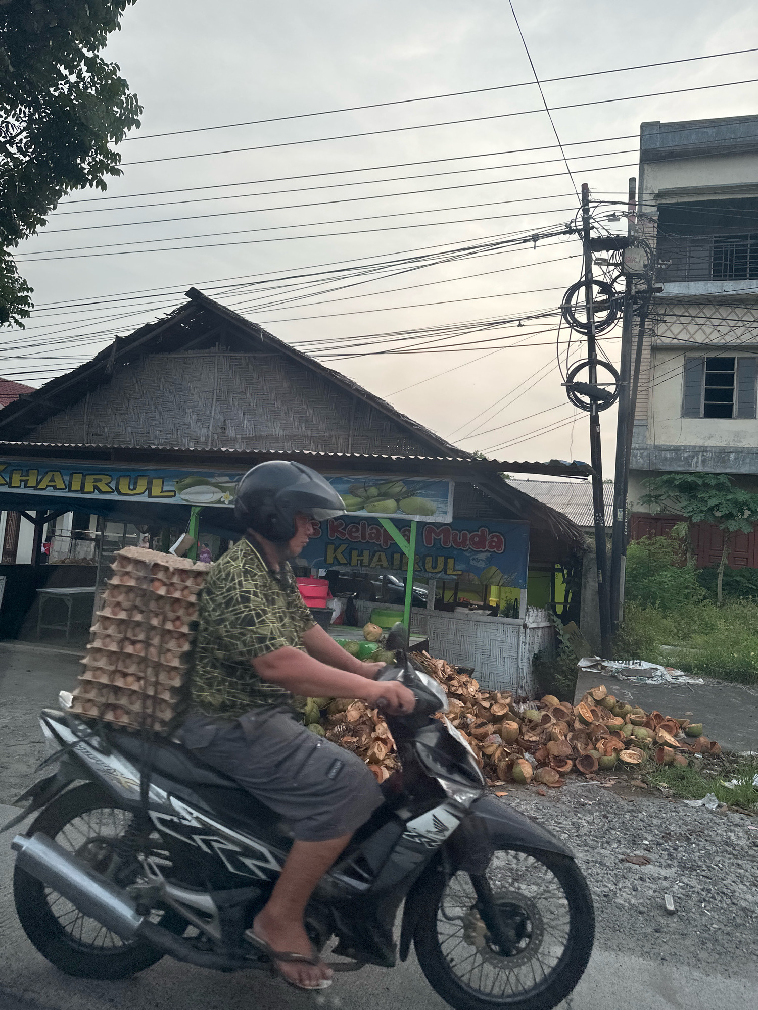 The image depicts a street scene in the City of Binjai, Indonesia, with a man riding a motorcycle carrying a large stack of egg cartons on his back. He is wearing a patterned shirt, grey shorts, a black helmet, and flip-flops. The motorcycle is black with white and grey accents.
In the background, there is a roadside stall with a thatched roof and bamboo walls. A sign above the stall reads "KHAIRUL" and "MUDA". The stall appears to be selling coconuts, as a large pile of halved coconuts is visible on the ground in front of it. There is also a building with multiple stories to the right of the stall, featuring windows and balconies. Overhead, numerous power lines crisscross the sky. The overall environment suggests a commercial area along a street.