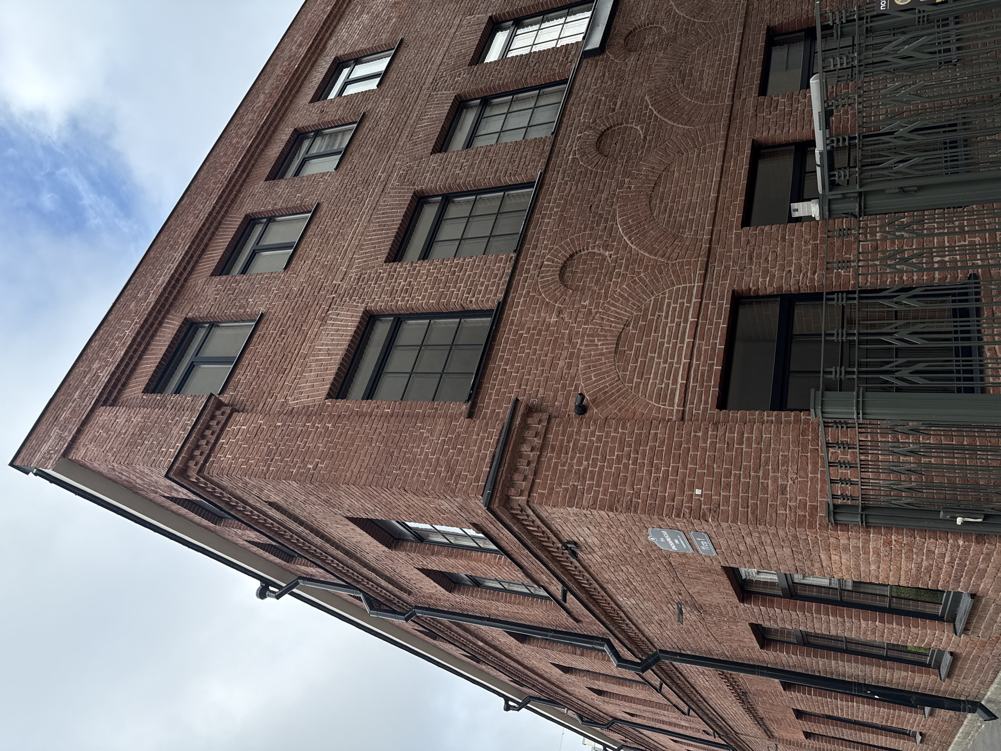 The image shows the corner of a red brick building in Saint Petersburg, Russia. The building has multiple windows with black frames and appears to be several stories tall. There are rain gutters on the side of the building and a black metal fence in front. Two street signs are visible on the building, one in Russian reading "Ивановская улица" (Ivanovskaya Street) and another with a building number. The sky is overcast with some hints of blue sky and clouds. The architectural style suggests it is an older building, possibly from the early 20th century or earlier, repurposed for modern use, perhaps as offices or loft apartments. There are no people or vehicles visible in the image. The overall scene is one of urban architecture under a cloudy sky.