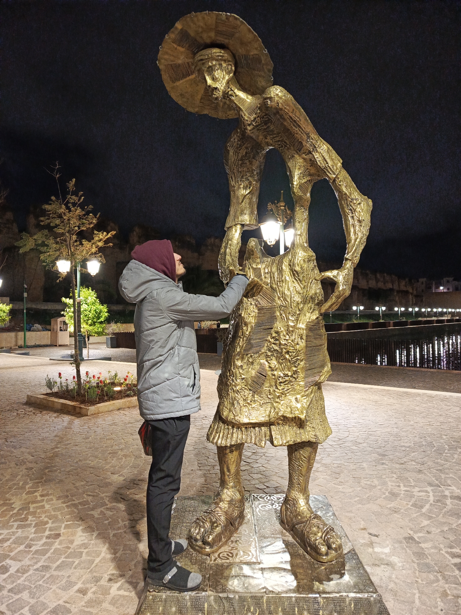 At night in the city of Al Machouar – Stinia, Morocco, a man wearing a grey jacket with a maroon hood, dark pants, and grey casual shoes stands on a patterned pavement beside a large golden statue of a man wearing a large hat. The statue holds an ornate streetlamp fixture in its arms. The man in the jacket is touching the statue with his right hand. In the background are trees, other streetlamps, and a body of water with lights reflected in it. A wall and buildings are visible beyond the water. The sky is dark.