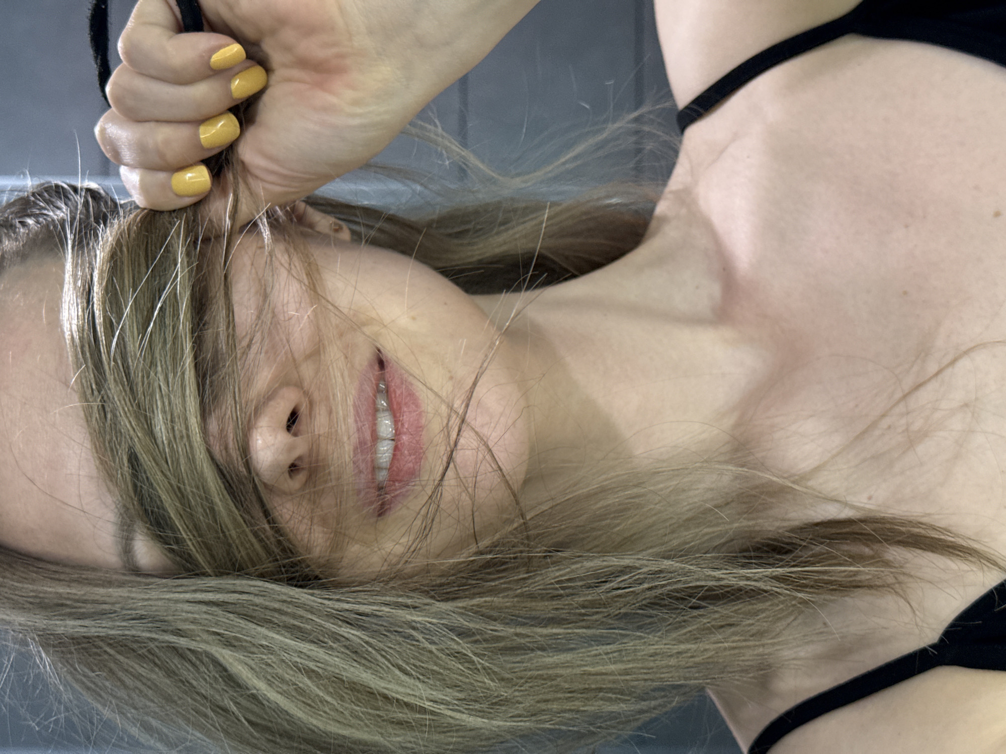 A woman lies on a gray surface. Her face is visible through strands of her light brown hair. She has yellow nail polish and is holding a black object. The location is in Chaikovsky, Russia.
