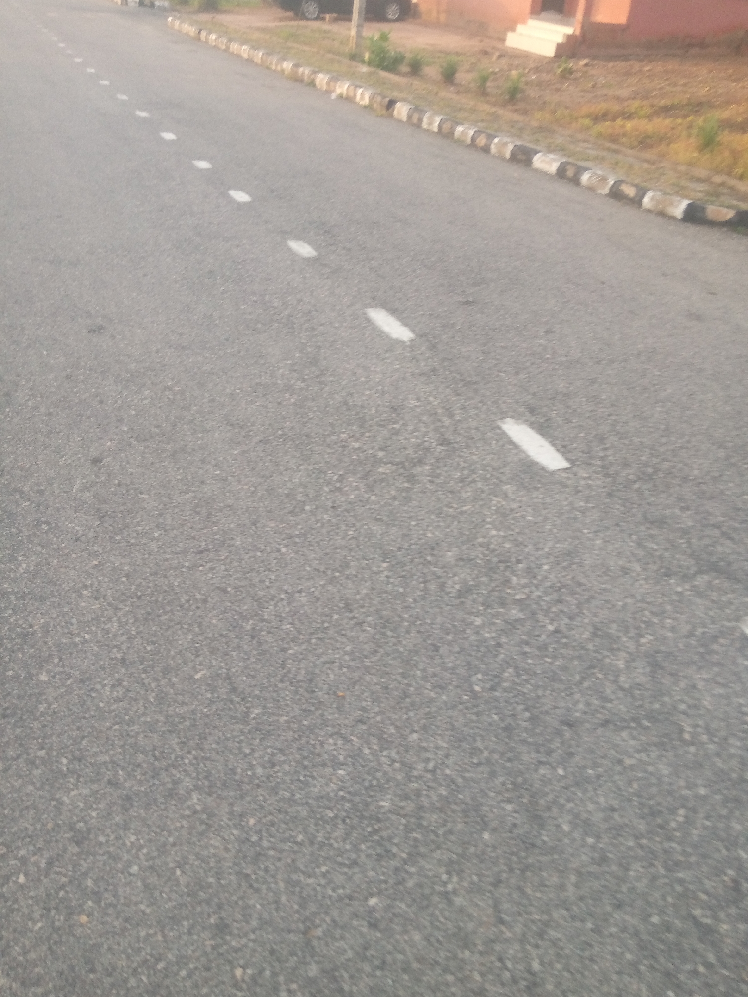 A straight asphalt road is visible with a series of white dashed lines marking the lane. To the right of the road, there's a border of alternating black and white painted concrete blocks. Beyond the border, dry grass and a building with light-colored steps can be seen. The sky is not visible in the frame. The image appears to be taken in Jauro Yinu, Nigeria.