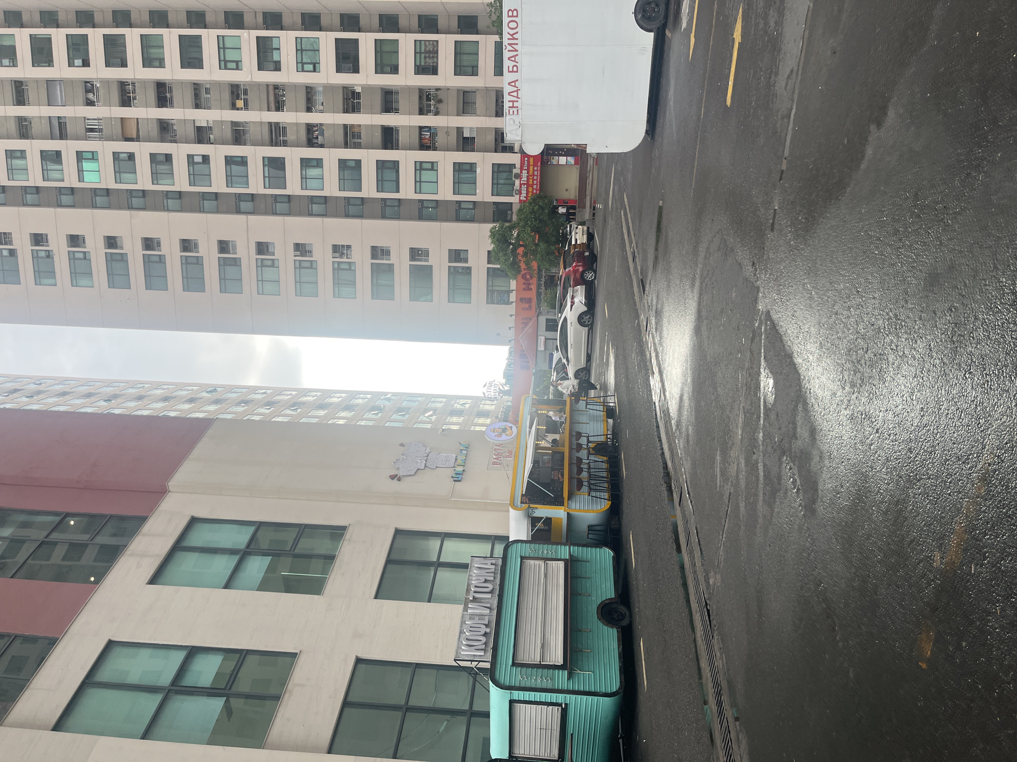 The image depicts a street scene in Bac Nha Trang Ward, Vietnam, on a wet day. A tall, modern building with many windows dominates the background. In the foreground, there are two food trucks/kiosks: one is turquoise and labeled "KOFE I TOCHKA!", the other is primarily white and yellow and appears to be a pasta bar. The street is wet and reflective, with visible yellow traffic arrows. Parked cars are visible between the buildings and the pasta bar. Signage for "Phuoc Thien Store" and "ARENDA BAIKOV" is also visible.