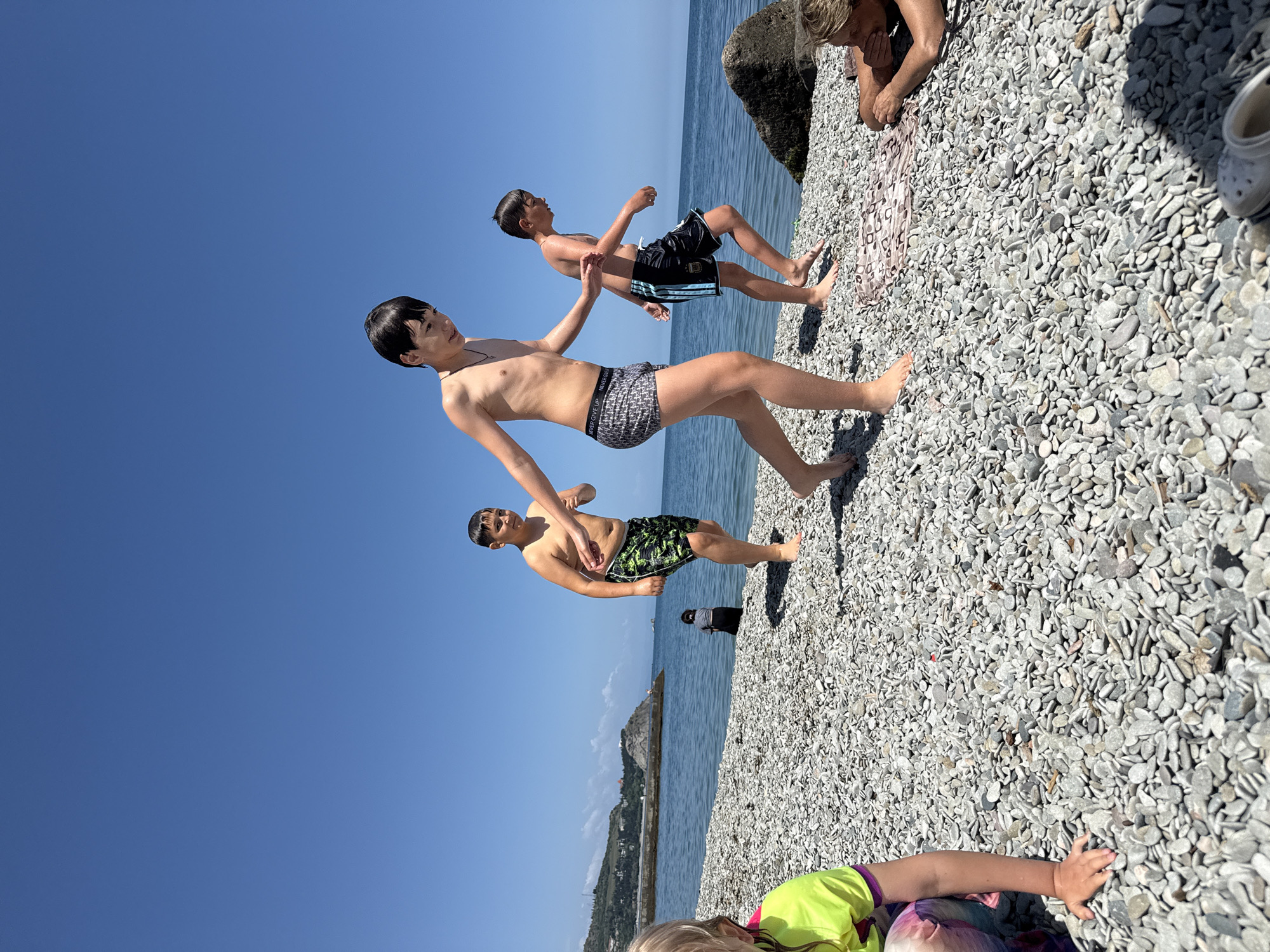 The image shows a group of people on a pebble beach. Three boys are standing and running near the water's edge. One boy is wearing gray and black patterned swim trunks, another has black and blue trunks, and the third has green and black trunks. Behind them is the ocean, and in the distance, a coastal town is visible with a mountainous backdrop. To the right, a person is lying on the pebbles with their head propped on their hands. At the bottom of the image, part of another person wearing a neon yellow and purple shirt is visible. Shoes are also near this person.