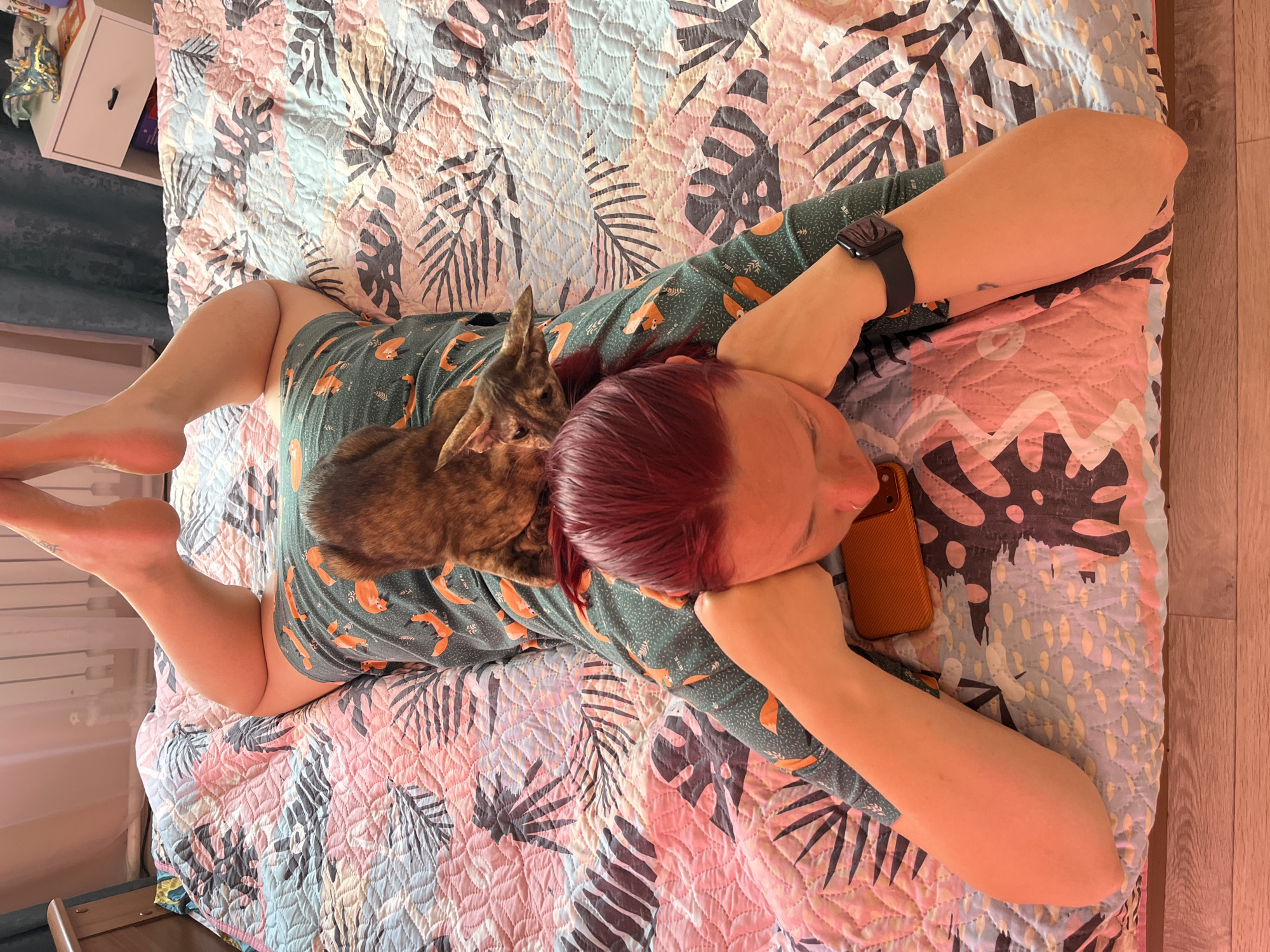 A high-angle, medium indoor shot depicts a Caucasian woman lying on her back on a bed with a cat on her chest. The woman has red hair and is wearing a green patterned pajama top. Her arms are bent, and her hands are near her face, holding what appears to be a smartphone with a burnt orange case. She is also wearing a black smartwatch on her left wrist. A brindle cat is lying on her chest, facing toward the woman's feet. The bed has a leaf-patterned quilt in shades of pink, blue, and black. A white cabinet with open storage space is visible in the upper left corner. The floor is made of light-colored wooden planks. The location is Chaikovsky, Russia.