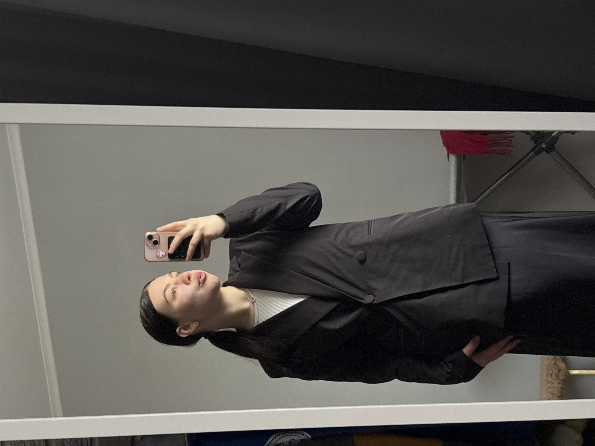 A young woman is taking a selfie in a mirror. She is wearing a black blazer over a white top and a black skirt. Her dark hair is pulled back in a ponytail. She is holding a pink and black smartphone. Behind her in the reflection is a gray wall and a black ceiling. A red scarf and the leg of a folding tripod are also visible. The reflection also shows a portion of the mirror frame. There is no verifiable information to confirm the location as Perm, Russia.