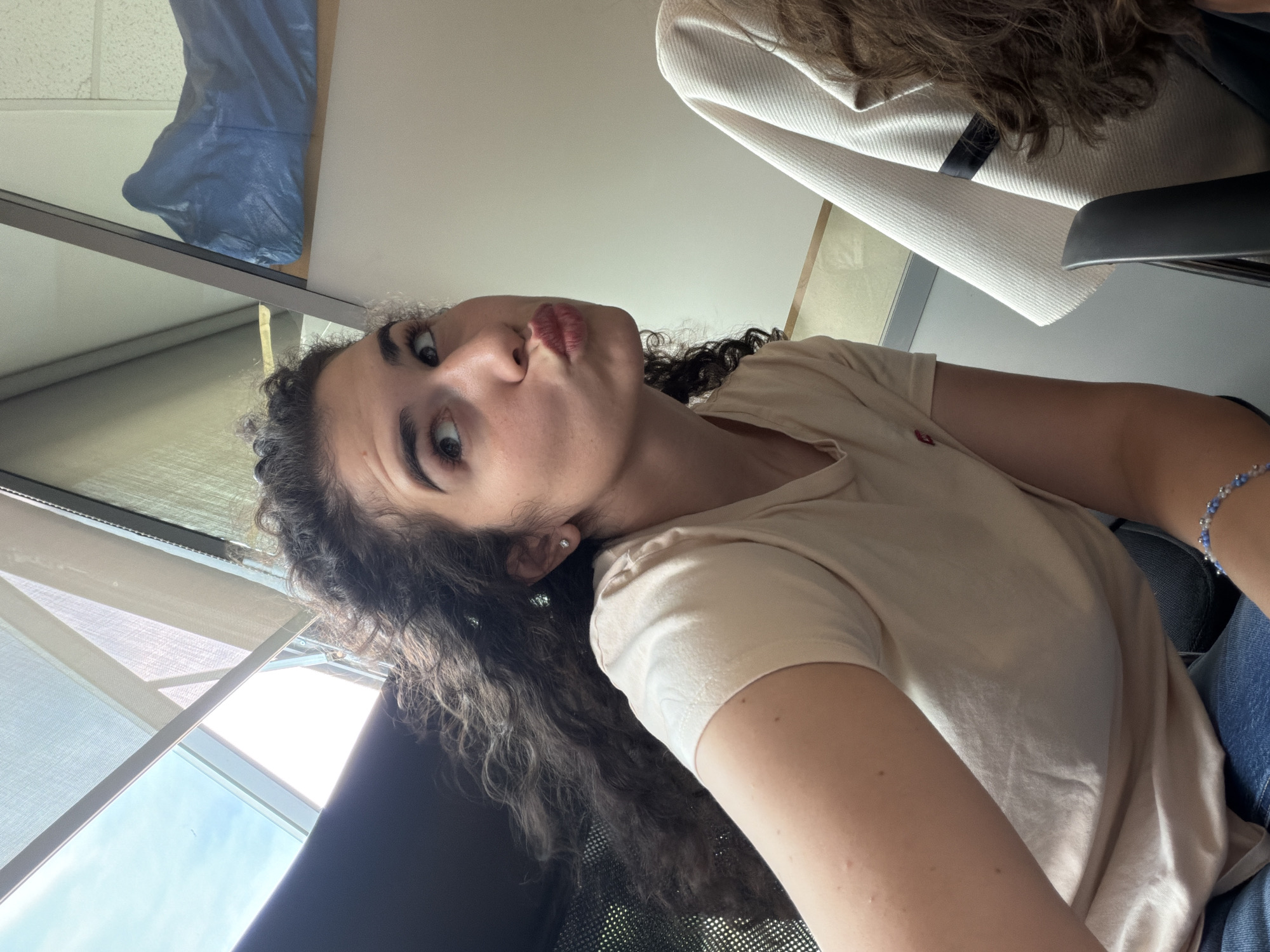 In Sofia, Bulgaria, a young woman with curly dark hair sits in a chair indoors, her body angled to the right. She wears a light-colored t-shirt and jeans, with a beaded bracelet on her left wrist. Her face is turned upward and to the left, her lips pursed in a playful expression, and her eyes looking in the same direction. Behind her, a window with blinds is visible, letting in natural light. To the right, a beige garment hangs, and a dark blue object is present on a shelf. The environment appears to be a room with plain walls.