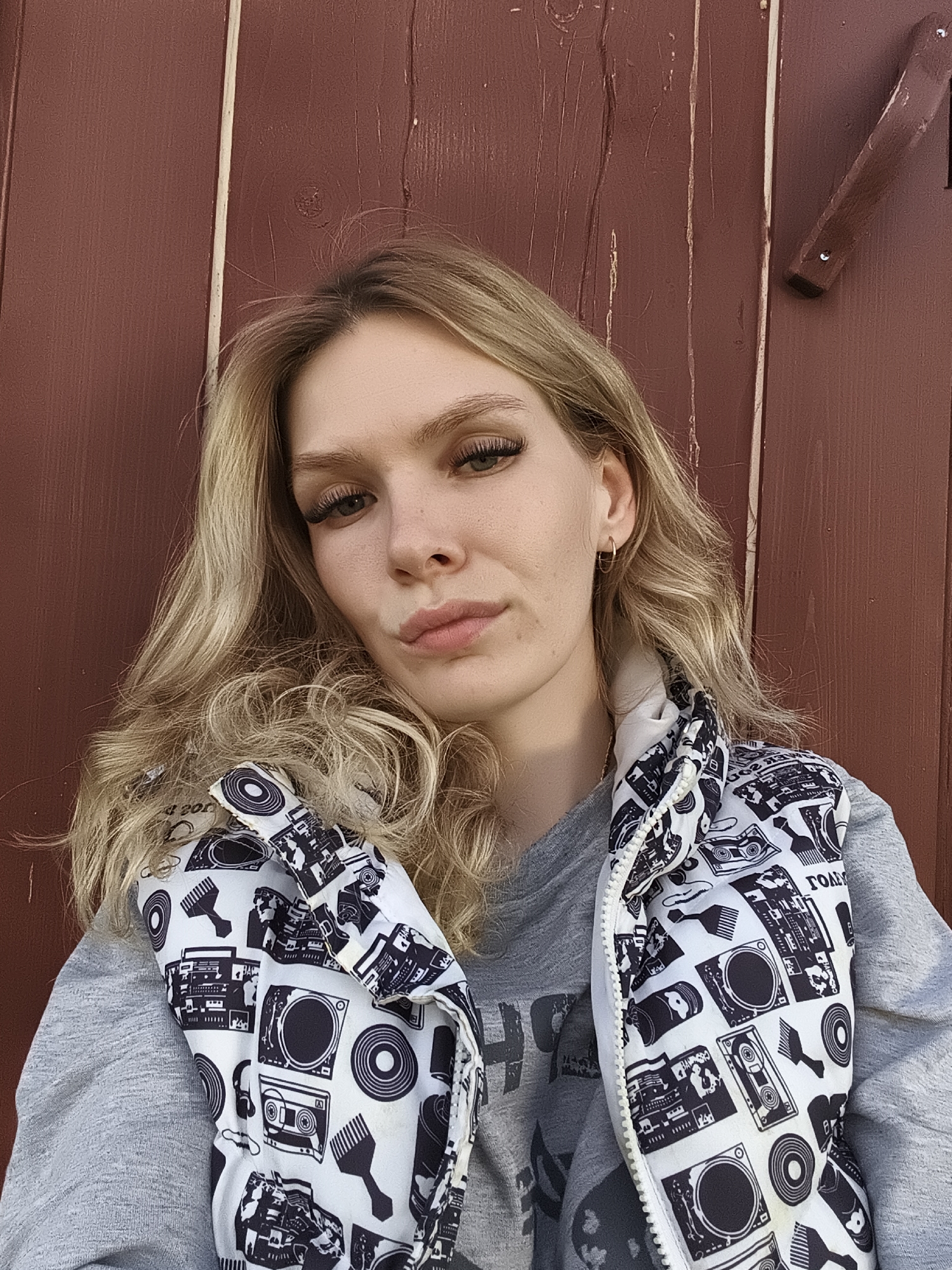 A young woman with long, wavy blonde hair is captured in a close-up selfie, posed against a dark reddish-brown wooden wall. Her light complexion is highlighted by soft, even lighting, suggesting daylight conditions. She looks directly at the camera with a neutral expression, lips slightly pursed. Her eyes are a hazel or green color, enhanced by long, dark eyelashes.
She wears a light grey long-sleeved top or sweatshirt, beneath a sleeveless puffer vest. The vest features a distinctive black and white pattern of various music-related items including turntables, boomboxes, cassette tapes, vinyl records, headphones, and what appear to be Afro picks. A white zipper runs down the front of the vest. A small gold hoop earring is visible in her right ear.
The background consists of vertical wooden planks, showing natural wood grain and knots, indicating an outdoor or rustic indoor setting, likely in Русский Вожой, Russia. To the right of her head, a dark wooden handle-like fixture is attached to the wall with two small fasteners. Some barely discernible dark text is visible on her grey shirt, obscured by the vest. On the patterned vest, the word "TOAM" (partially visible) and "d 200" can be seen among the design elements.