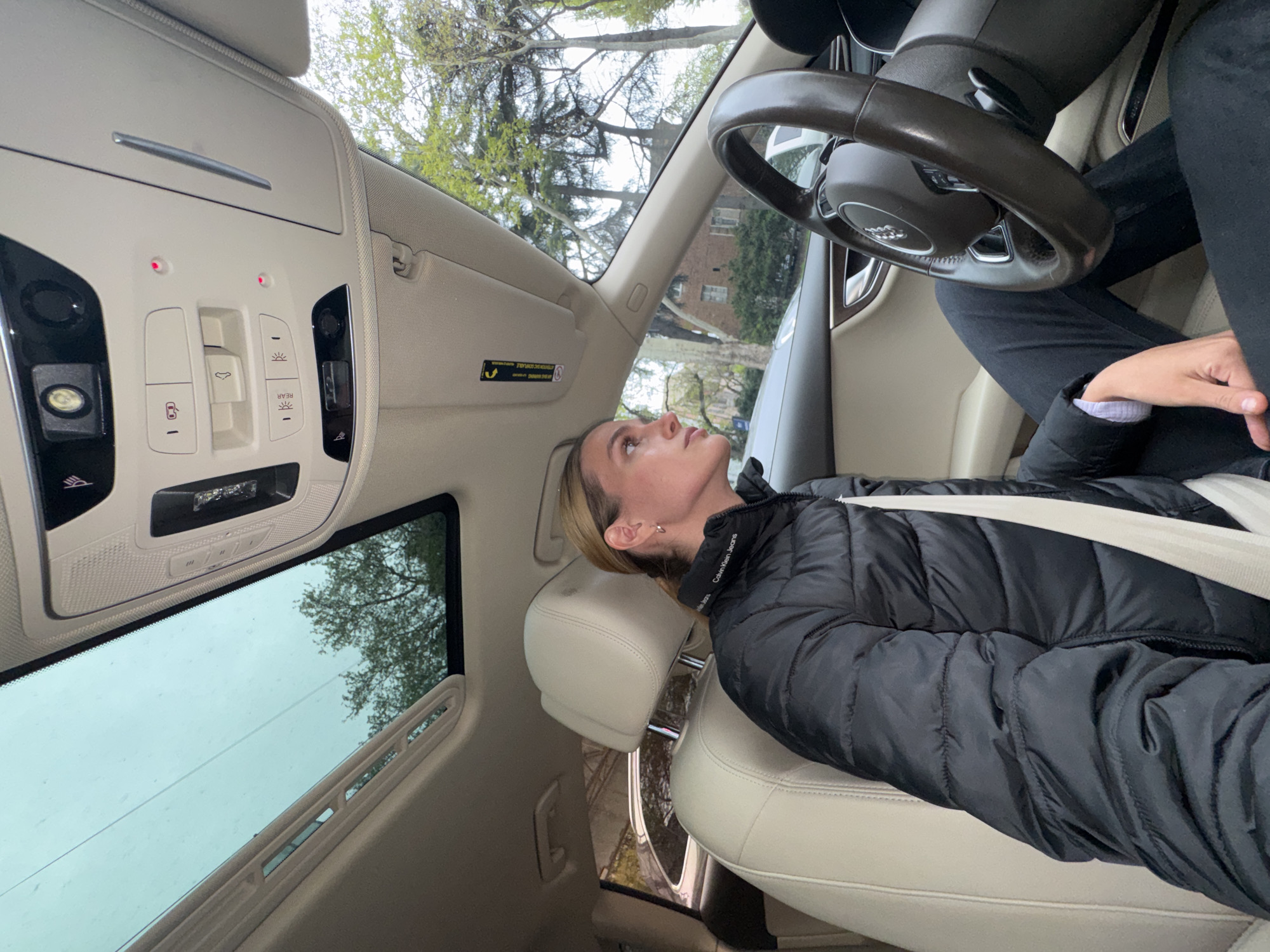A woman with light hair is seated in the driver's seat of a stationary Audi car, looking upwards with a neutral expression. She is wearing a black puffer jacket with "Calvin Klein Jeans" visible on the collar and has a light-colored seatbelt across her chest. The car interior features luxurious beige leather seats and a dark brown steering wheel with the Audi logo.
Above her, the car's overhead console is visible, showing several buttons and controls. Two small red indicator lights are illuminated. Buttons include controls for interior lights, a sunroof, and one labeled "REAR" for rear lighting. A sun visor with warning labels is also present. The car is equipped with a panoramic sunroof, through which a light grey, overcast sky and trees with green foliage are visible. Raindrops are present on the sunroof glass, suggesting recent precipitation or an ongoing light rain. Through the windshield, beyond the trees, a building can be vaguely seen in the background. The scene is set outdoors during daylight in Tbilisi, Georgia.