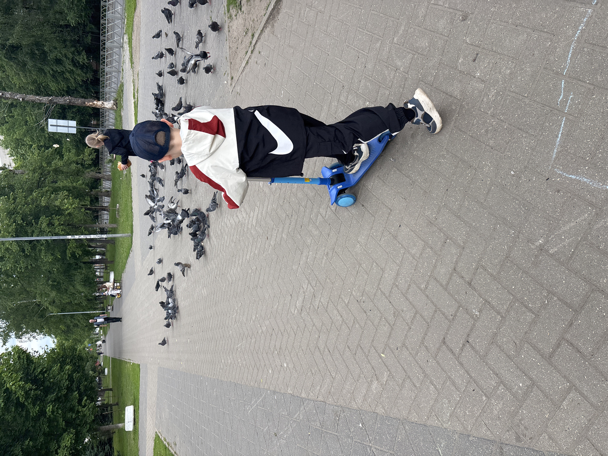 The image depicts an outdoor scene in a park or pedestrian area in Rusanovo, Russia, during daytime. The primary subject is a child, seen from behind, wearing a dark blue baseball cap, a white and maroon jacket, and black pants with a prominent white Nike swoosh logo on the left leg. The child stands on a blue three-wheeled scooter.
Numerous pigeons are gathered on the light grey paved path, which features a herringbone brick pattern. A woman with blond hair, wearing a black jacket, is partially visible beyond the child, holding what appears to be a piece of bread, suggesting she is feeding the birds. The child appears to be observing or interacting with the pigeons.
The paved pathway is bordered by green grass and lush trees. In the background, another person is walking further down the path, and a white stroller or cart is discernible in the distance. A signpost is also visible in the middle ground, though its details are indistinct. A faint white chalk line is present on the pavement to the right. The scene conveys a calm, leisurely activity in a public green space. The only visible text is the Nike logo on the child's pants.