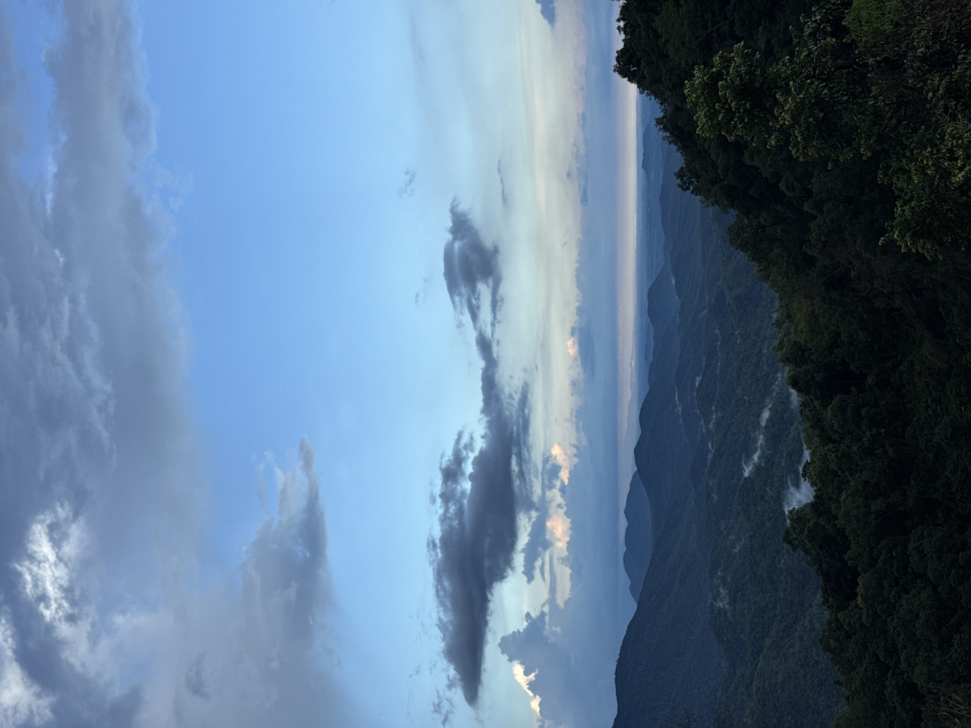 This image depicts a sweeping mountain landscape under a partly cloudy sky. The foreground is dominated by dark, densely packed trees, suggesting a forest-covered mountain slope. Beyond this, a series of rolling, verdant mountains recede into the distance, with layers of mist or low-lying clouds settling in the valleys between them. The sky is a mix of bright blue and darker, diffused clouds, with some hints of pink or orange light suggesting either dawn or dusk. The overall impression is one of a vast, natural environment, possibly a national park or a scenic overlook. There are no people or man-made structures visible in this image. The location is stated to be in Nao Sẻ, Vietnam, a region known for its mountainous terrain and natural beauty.