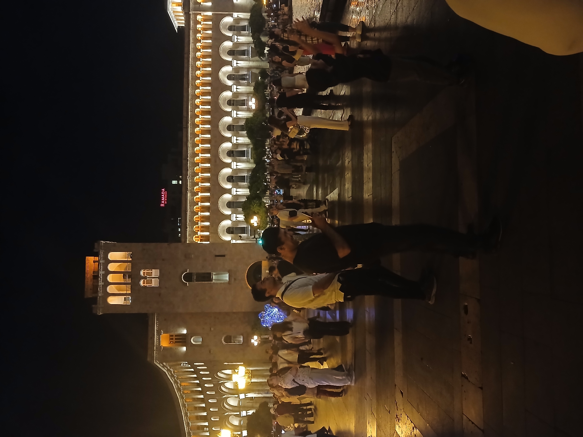 A nighttime, exterior, wide shot captures a public square with people gathered in front of a grand, illuminated building. The building, constructed of stone, features a tall, central tower with arched windows, and a long, colonnaded facade. The architecture is illuminated by warm, yellow lights, highlighting arches and decorative elements. A red "Ramada" sign is visible on the upper portion of the building.
A crowd of people, diverse in age and gender, are standing and walking in the square. They are dressed in casual attire. One man in the foreground, wearing a black t-shirt, is looking at his phone. Another man beside him, in a white polo shirt, stands looking forward. A woman in a white top and light-colored pants is also visible. Some individuals are in small groups, while others are walking alone. A bright blue, illuminated decorative object, possibly a sculpture or light installation, is present in the mid-ground. The ground surface appears to be paved with stone or tiles, and some areas show reflections, suggesting it may be wet. Trees are sparsely planted along the edge of the building. The scene is illuminated by the building's lights and street lamps, creating a somewhat dim and atmospheric environment.