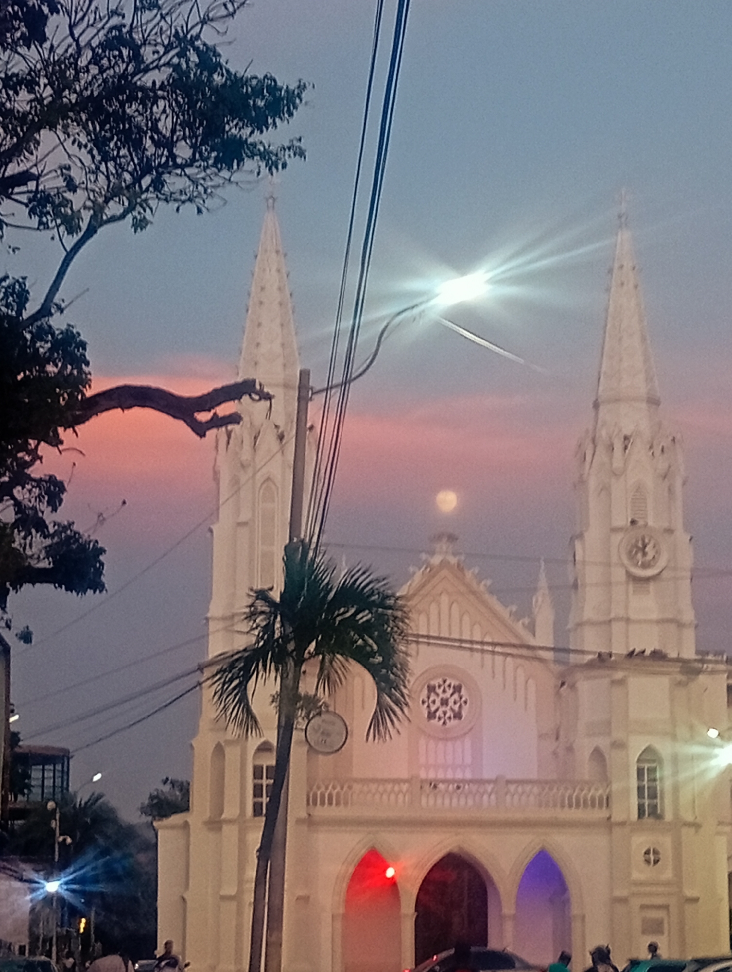 The image displays an evening scene of a prominent white church in Juan Griego, Venezuela, set against a twilight sky painted with pink, orange, and blue gradients. The church, exhibiting Gothic Revival architecture, features two ornate spires, each topped with a cross, and a circular rose window on its central facade. A clock face is visible on the upper section of the right spire. The lower arches of the church are illuminated with distinct red and purple/blue lights, adding a dramatic flair. A faint moon is visible in the sky above the church's main structure.
In the foreground, dark tree branches, including a large tree on the left and a prominent palm tree near the center, partially frame the church. Numerous power lines crisscross the sky. Several bright street lights create starburst lens flares, indicating the low-light conditions. At street level, several indistinct figures of people are visible, some appearing to be standing or walking, while the tops of vehicles are also present. One individual in the mid-ground appears to be holding a musical instrument. An indistinct round sign hangs on the right side of the church building. The overall scene evokes a peaceful and atmospheric evening in Juan Griego.