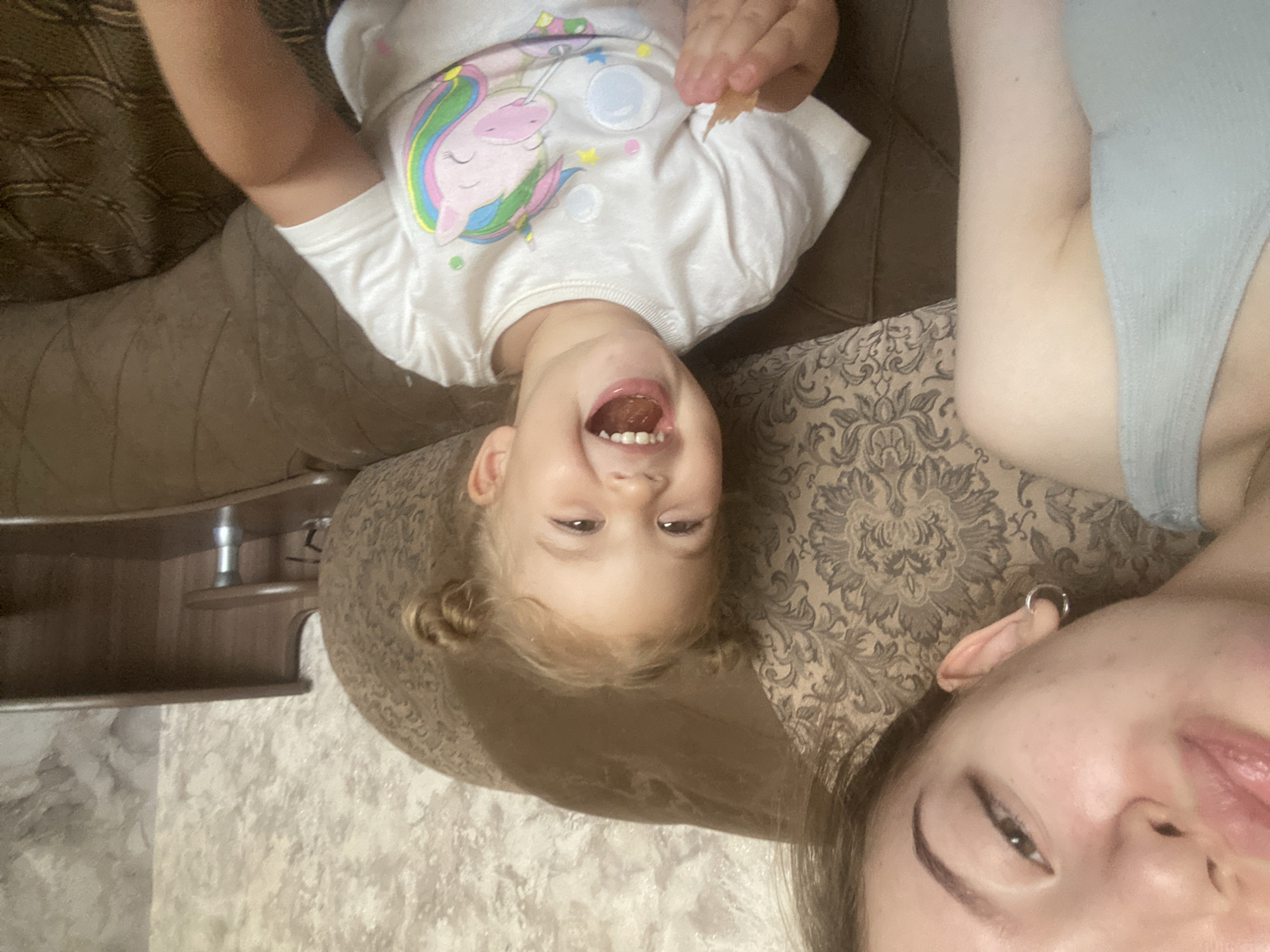 The image is a selfie taken by a young woman who is partially visible on the left side of the frame, leaning in towards the camera. She is wearing a light blue, sleeveless top. Her lips are slightly parted, and she is looking off to the side. A silver hoop earring is visible in her left ear. In the center and right of the frame is a young child, likely a girl, with blonde hair styled in two pigtails. She is wearing a white t-shirt with a colorful unicorn graphic. The child is laughing, with her mouth wide open and teeth visible. She is holding a small piece of food in her right hand. The two subjects are seated on a large, brown, upholstered couch with a patterned fabric. In the background, there is a textured wall and a wooden piece of furniture that appears to be a desk or shelf unit. The overall scene appears to be indoors, likely a home environment. The location is stated to be in Pesnevo, Russia.