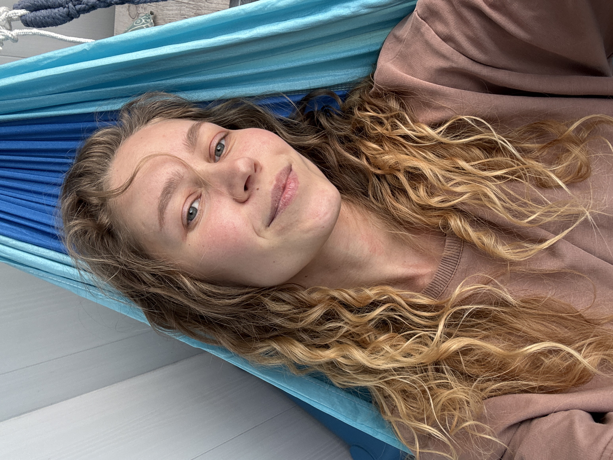 A woman with long, curly blonde hair is lying in a blue hammock. She is wearing a brown sweatshirt. The hammock is suspended near a light-colored wooden wall or surface. The location is unknown; there is no evidence to suggest it is in Kladovitsy, Russia.