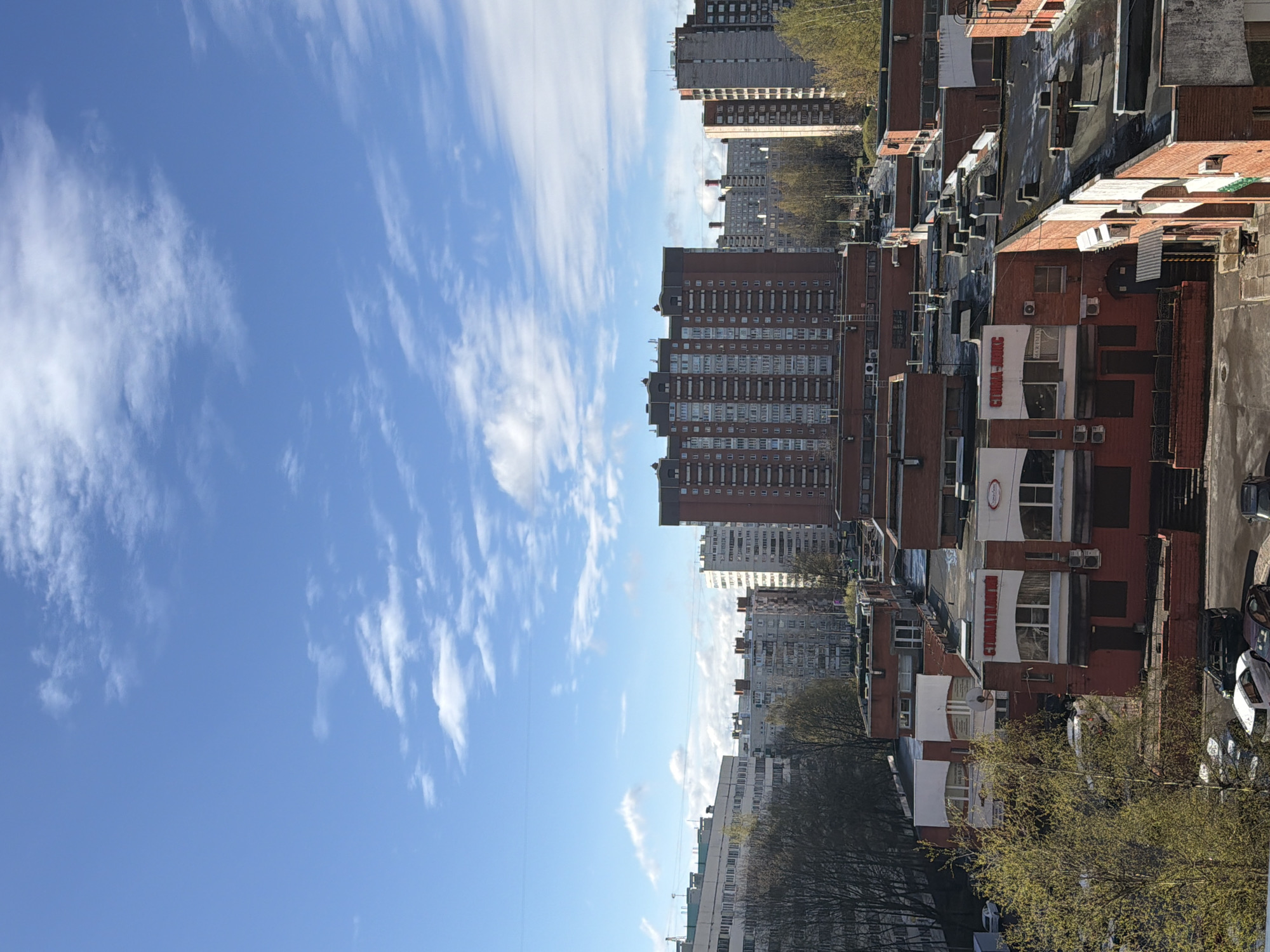 The image, correctly oriented, displays a bright, sunny urban landscape in Saint Petersburg, Russia. The left side of the frame is dominated by a clear blue sky with scattered, wispy white clouds, indicating fair weather.
The right side and lower portions of the image show a dense arrangement of buildings. In the background, several tall, multi-story buildings, likely residential or commercial, stand against the sky, characterized by numerous windows. In the mid-ground and foreground, a series of lower-rise buildings with red-brick facades and a mix of flat and pitched roofs are visible. These buildings feature multiple windows, air conditioning units, and satellite dishes.
Trees with budding green leaves, suggesting early spring, are interspersed among the buildings and line a street or park area in the lower left. Several cars are parked on a paved surface at the bottom right.
Visible text in Cyrillic script appears on the facades of the red-brick buildings. A prominent red sign with white letters clearly reads "СТОМА-ИДЕАЛ" (STOMA-IDEAL). Below it, another sign states "МЕДИЦИНСКИЙ ЦЕНТР" (MEDICAL CENTER), and a partially obscured sign further down appears to be "ОРТОПЕДИЯ" (ORTHOPEDICS). A white circular logo with red text, likely associated with "STOMA-IDEAL," is also present. The scene is illuminated by bright daylight, suggesting morning or early afternoon.