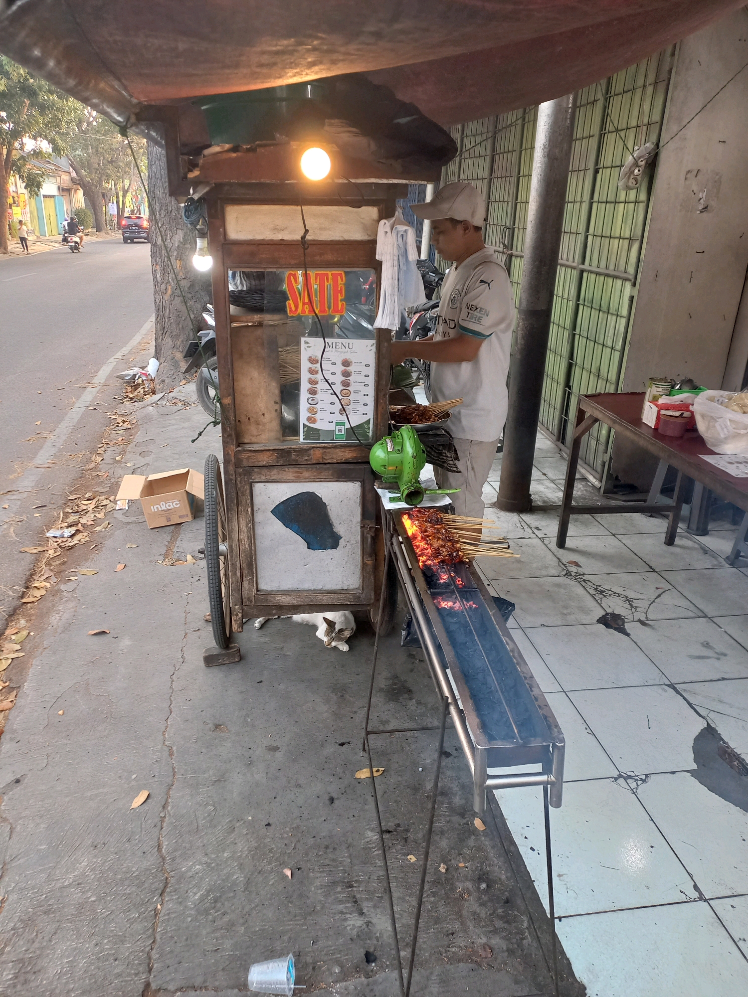 A street vendor is grilling satay in Cimahi, Indonesia. The vendor, a man wearing a white cap and a t-shirt, is operating a portable satay cart. The cart has a grill with burning charcoal where several skewers of satay are being cooked. Behind the vendor, a menu board displays various satay options. A green fan is positioned near the grill. The cart is situated on a sidewalk next to a street with passing vehicles. In the foreground, a cat is resting underneath the cart. A cardboard box with the logo "inlac" is visible on the sidewalk. The environment appears to be an outdoor urban setting with trees and buildings lining the street. The time of day seems to be late afternoon or early evening, given the lighting conditions.