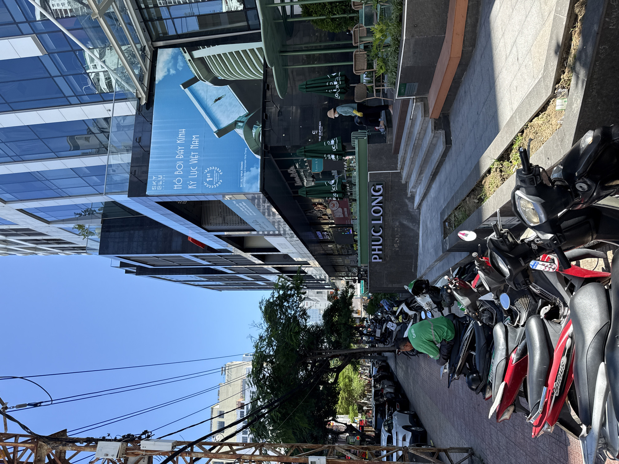 The image shows a street scene in Nha Trang, Vietnam, on a clear, sunny day. A modern, multi-story building with a glass facade dominates the right side of the frame. A large blue billboard on the building advertises "SKY BLU LOUNGE" with the text "HỒ BƠI ĐÂY KÍNH KỶ LỤC VIỆT NAM" which translates to "Glass bottom pool Vietnam record". Below this, it mentions "SKY BLU NHA TRANG" and "THE EMPYREAN NHA TRANG".
In the foreground, a row of parked motorcycles lines the sidewalk. One person, wearing a green shirt with a "Grab" logo, sits on a motorcycle. Further down the sidewalk, a person in a light-colored hat and green top, wearing yellow gloves, is walking up a set of outdoor stairs leading to an entrance area. This area features outdoor seating with green umbrellas and chairs. A sign on the building reads "PHUC LONG".
The left side of the image features a utility pole with numerous electrical wires. A line of cars is parked along the street, and trees line the sidewalk. The overall environment suggests an urban setting with commercial establishments and residential buildings.