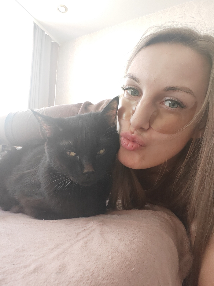 This image depicts a female subject interacting with a black cat in an indoor setting within the city of Chaikovsky, Russia. The woman, positioned on the right side of the frame, has light brown hair and green eyes, with translucent gel patches applied beneath her eyes. Her lips are pursed in a kissing gesture, directed towards the camera. A solid black cat is positioned to her left, resting on a light pink or beige textured surface. The cat's head is turned towards the camera, displaying green-yellow eyes and a neutral to slightly discontented expression. The background is overexposed, revealing a window with light-colored curtains on the left and a white wall, indicating a bright ambient light source. A circular recessed light fixture is visible on the ceiling in the upper-center background.