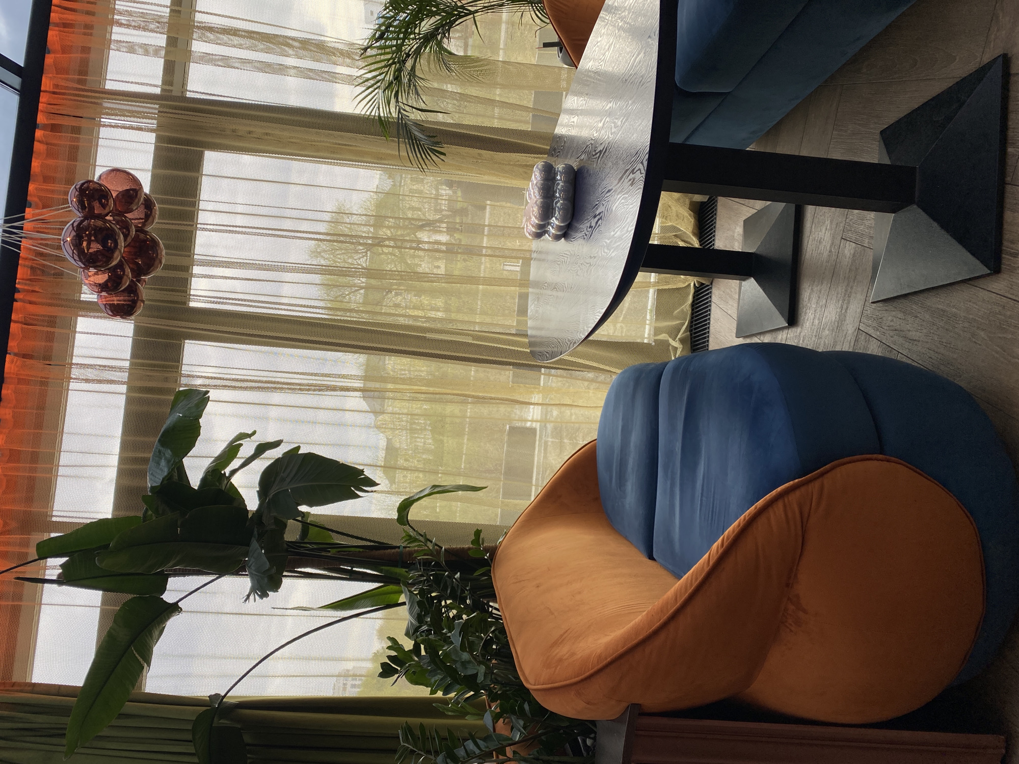 The image depicts the interior of a room, likely a restaurant or cafe, with a focus on the decor and furniture. A round, dark-colored table with a unique pedestal base is visible. On the table, there is a decorative object resembling a row of spheres. A blue velvet sofa or booth runs alongside the table.
In the foreground, an orange velvet chair with a blue cushion is featured. Plants, including a large-leafed one, are placed near the window, adding a touch of greenery to the space.
The window is covered by sheer, light-filtering curtains. Hanging near the window is a decorative piece comprised of interconnected spheres.
Based on online information, the furniture and decor style, it is likely the location is in the city of Yekaterinburg, Russia. There is no evidence of people.