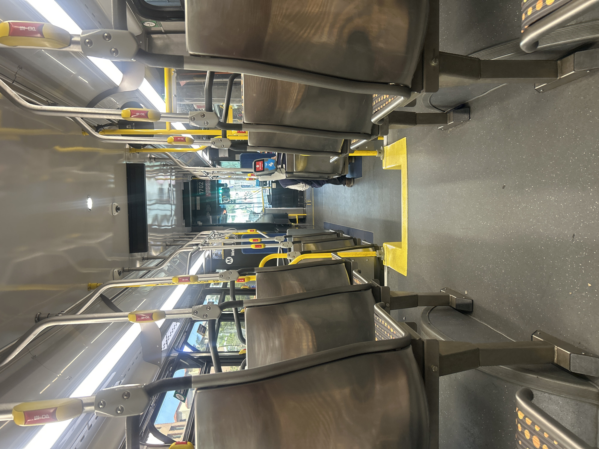The image depicts the interior of a Los Angeles Metro bus, looking towards the front. The bus is mostly empty, with rows of grey seats visible on either side of the aisle. Yellow poles and handholds are present throughout the interior, aiding passengers in maintaining balance while the bus is in motion. At the front of the bus, the driver's area is visible, with a digital display showing "1702". The bus is likely stopped, as the view through the front windshield reveals urban scenery with buildings and trees. A person is visible near the fare payment kiosk. The floor is dark and textured, likely for slip resistance. Overall, the scene conveys a typical public transportation environment in Los Angeles.