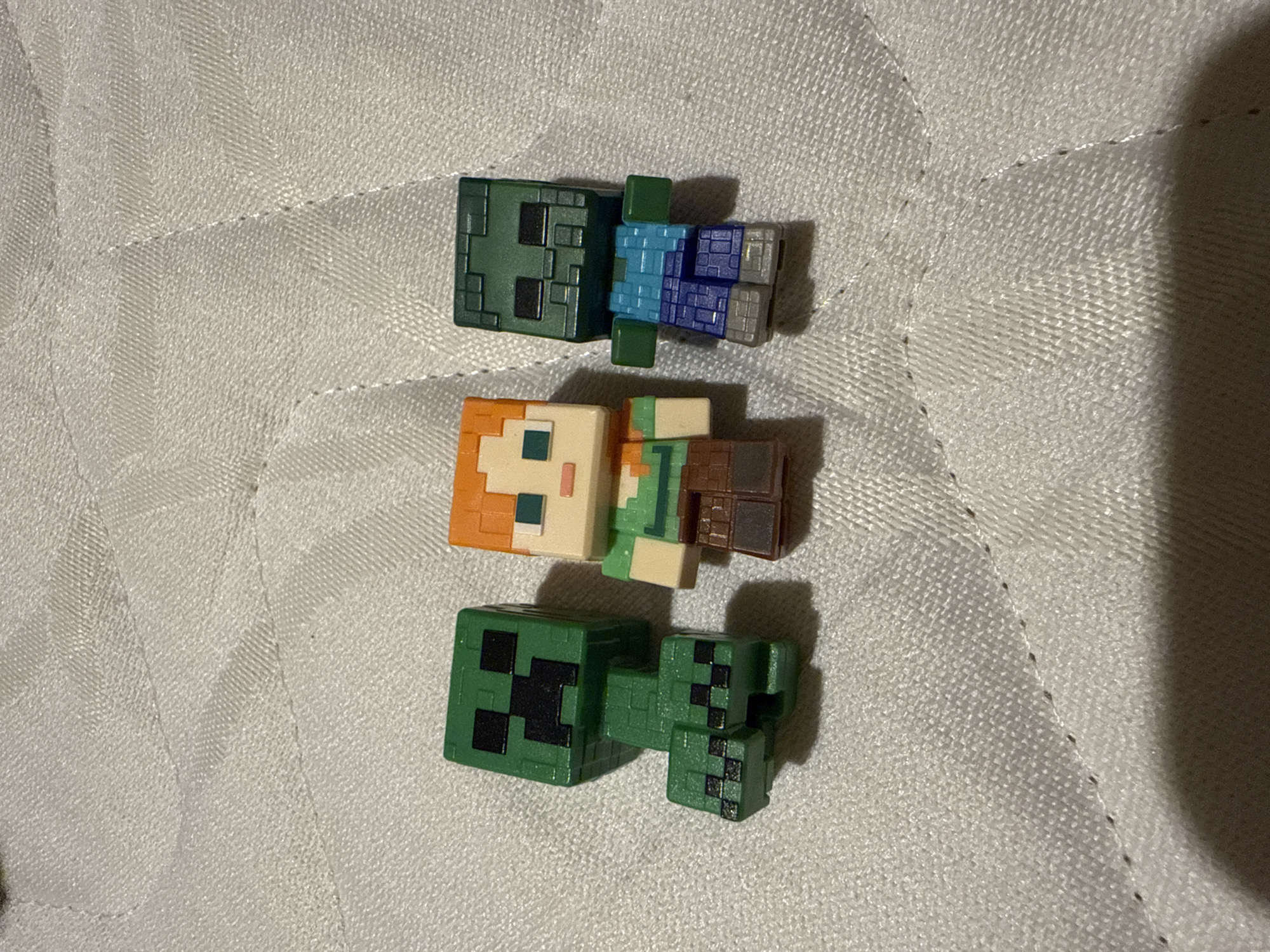 The image features three small, block-style plastic figurines, characteristic of characters from the video game Minecraft, arranged vertically on a light-colored, textured fabric surface. The surface appears to be a mattress or similar upholstered material, with subtle stitched lines forming a diagonal pattern.
From top to bottom, the figurines are:
1. A Zombie character, depicted with a dark green head and torso, light blue sleeves, dark blue pants, and grey feet. Black pixelated details mark its eyes and mouth.
2. An Alex character, identifiable by her orange-brown hair, light skin, a light green shirt, and brown pants or a backpack. A single blue pixel represents one eye.
3. A Creeper character, entirely green with its signature black pixelated facial features on its cube-shaped head.
The figurines are stationary, simply laid out on the fabric. The lighting suggests an indoor setting, likely under artificial light, as soft shadows are visible to the bottom right of each figure. There are no discernible time-of-day indicators, weather elements, or visible emotions. No text is present in the image. The location context provided is Sofia, Bulgaria.