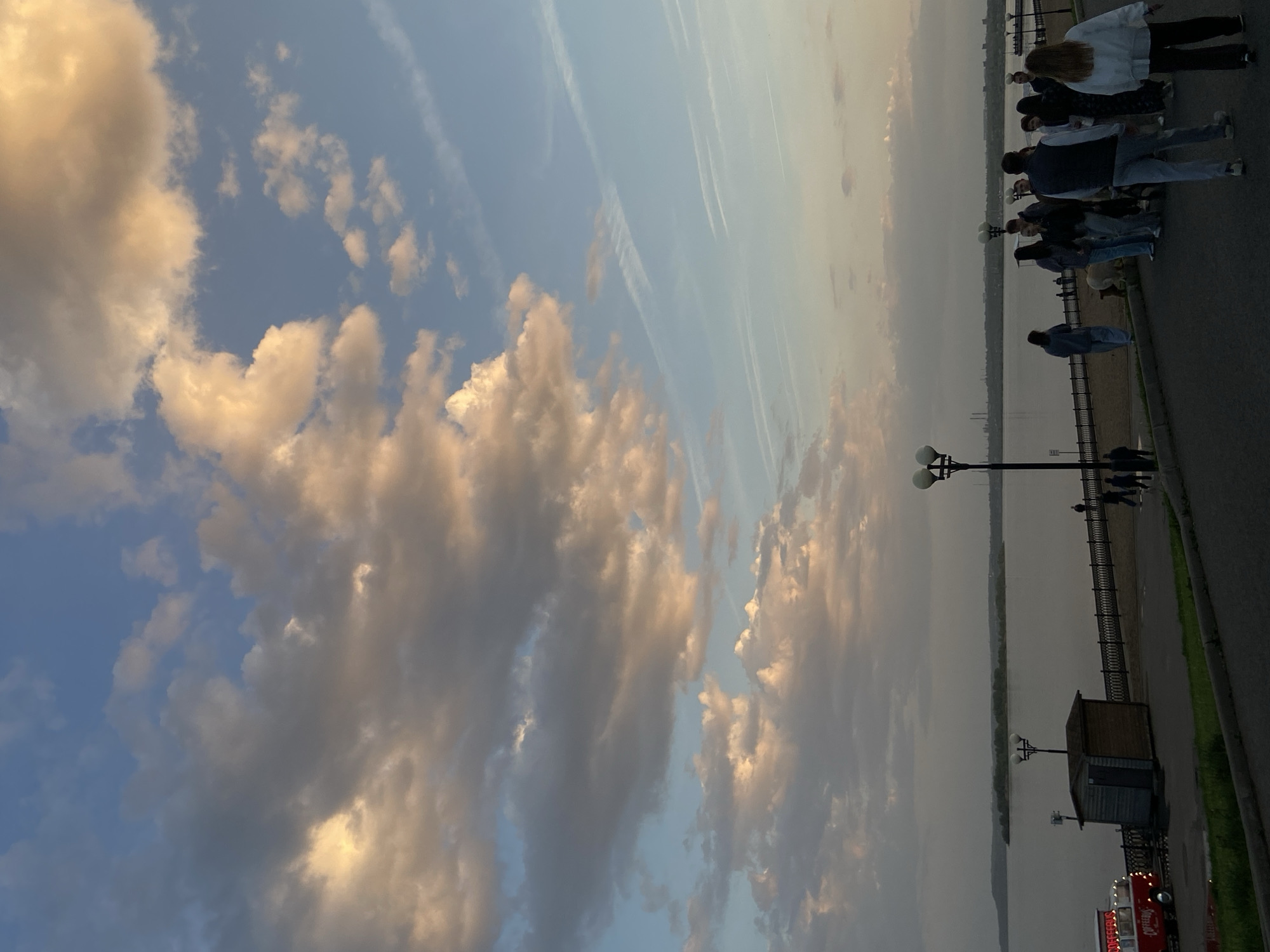 The image captures an outdoor scene in Saratov, Russia, near the Volga River. The sky is filled with clouds, some illuminated by the setting sun. Contrails from aircraft are also visible. Along the riverbank, a group of people is walking on a paved path next to a metal railing. Street lamps are spaced along the path. A small structure, possibly a shed, and a food truck are present near the river. The river surface is calm.