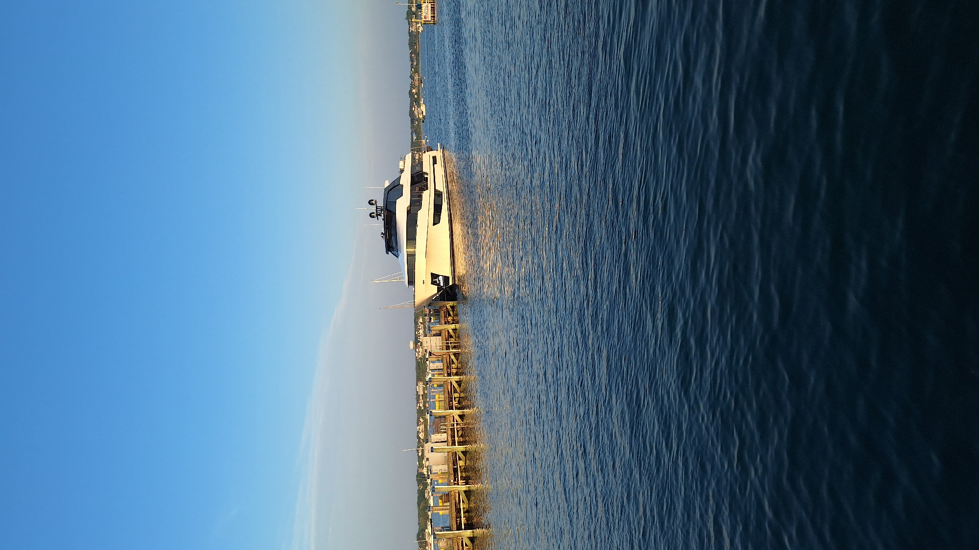 A white luxury yacht is docked at a wooden pier in Nantucket, USA. The yacht is positioned to the right of the frame, its sleek hull reflecting the sunlight. The pier extends from the left side of the image, with several other boats visible in the distance. The water is calm, with gentle ripples reflecting the clear blue sky. The background shows a line of houses and trees, suggesting a coastal town. The lighting indicates it is likely late afternoon or early evening, with the sun casting a warm glow on the scene. No people are visible in the image.