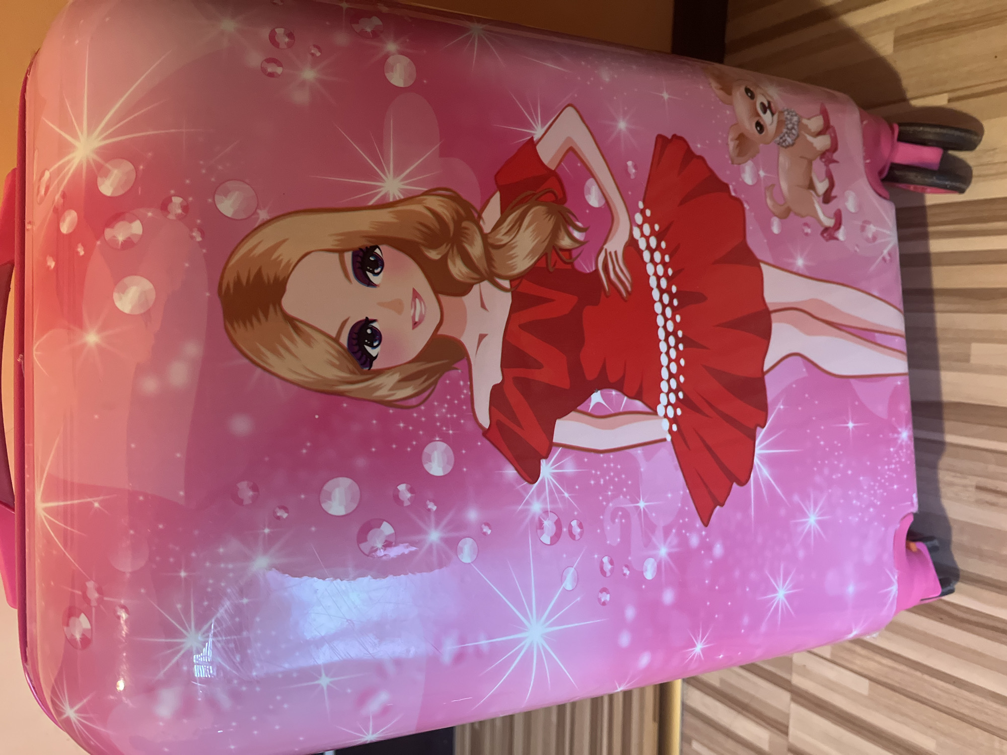 The image shows a pink, hard-sided suitcase decorated with a cartoon image of a blonde woman in a red dress and a small dog. The suitcase has a handle on one side and wheels at the opposite end. The background is a blurry view of a wooden floor. There is no information in the image to suggest this is in Burgas, Bulgaria.