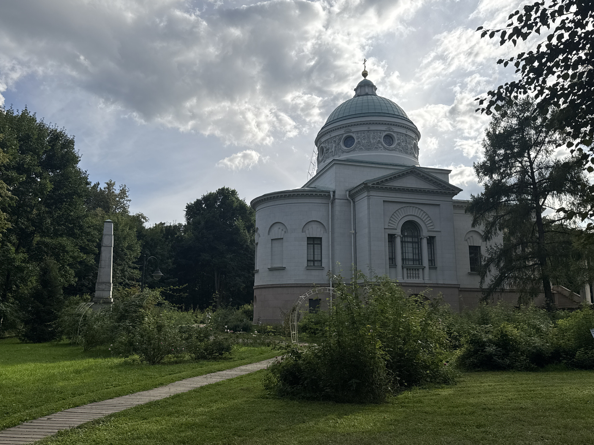 A light gray, neoclassical building with a green dome and a cross on top stands in a park. The building features arched windows and decorative elements. In the foreground, a wooden pathway winds through a grassy area with shrubs and trees. To the left of the building, a tall, narrow monument stands in the distance. The sky is partly cloudy, with sunlight breaking through the clouds. The scene appears to be in a quiet, historical park setting, likely the estate in Arkhangelskoye.